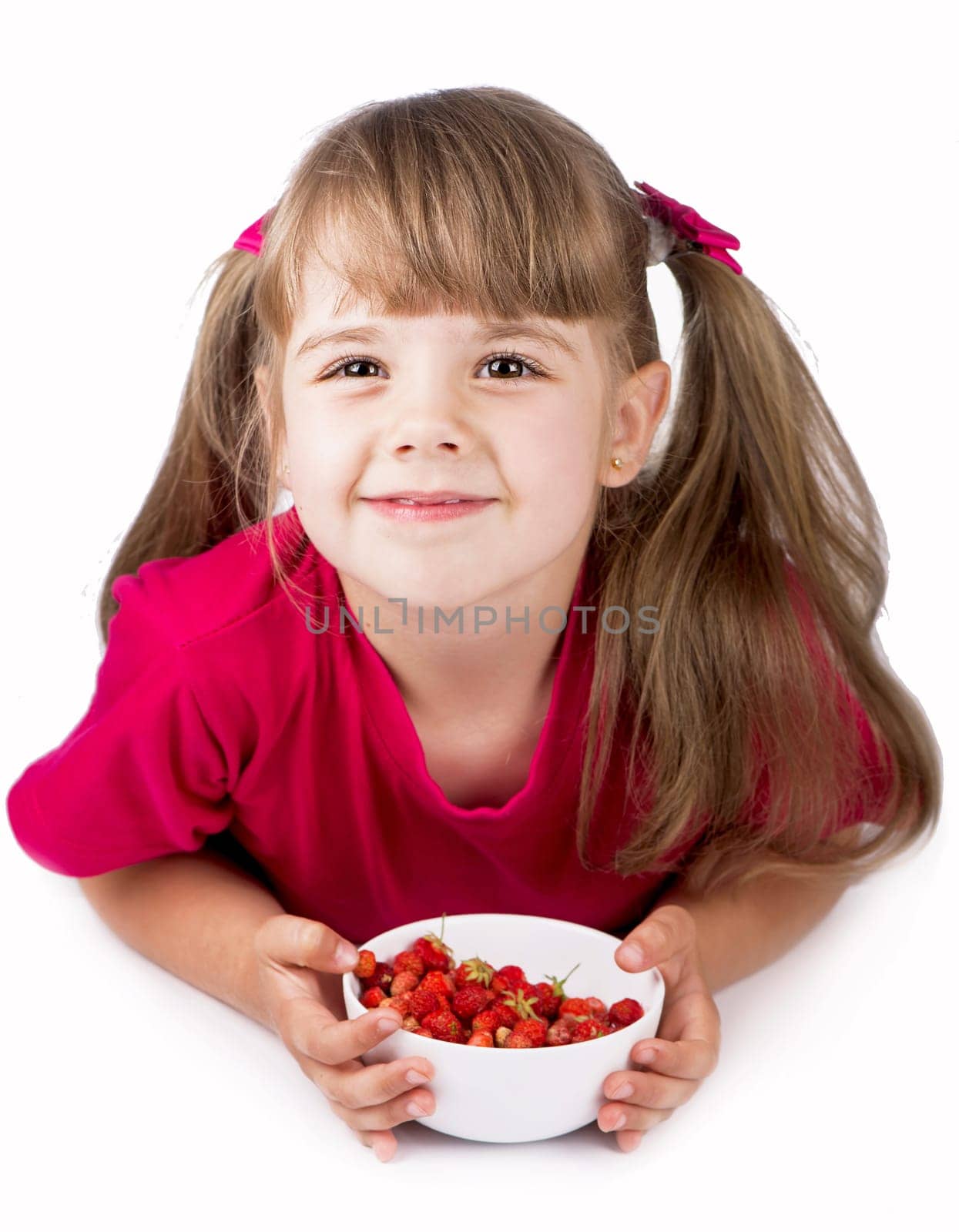 Portrait of a cheerful little girl in a pink t-shirt holding a white bowl of strawberries on a white background by aprilphoto