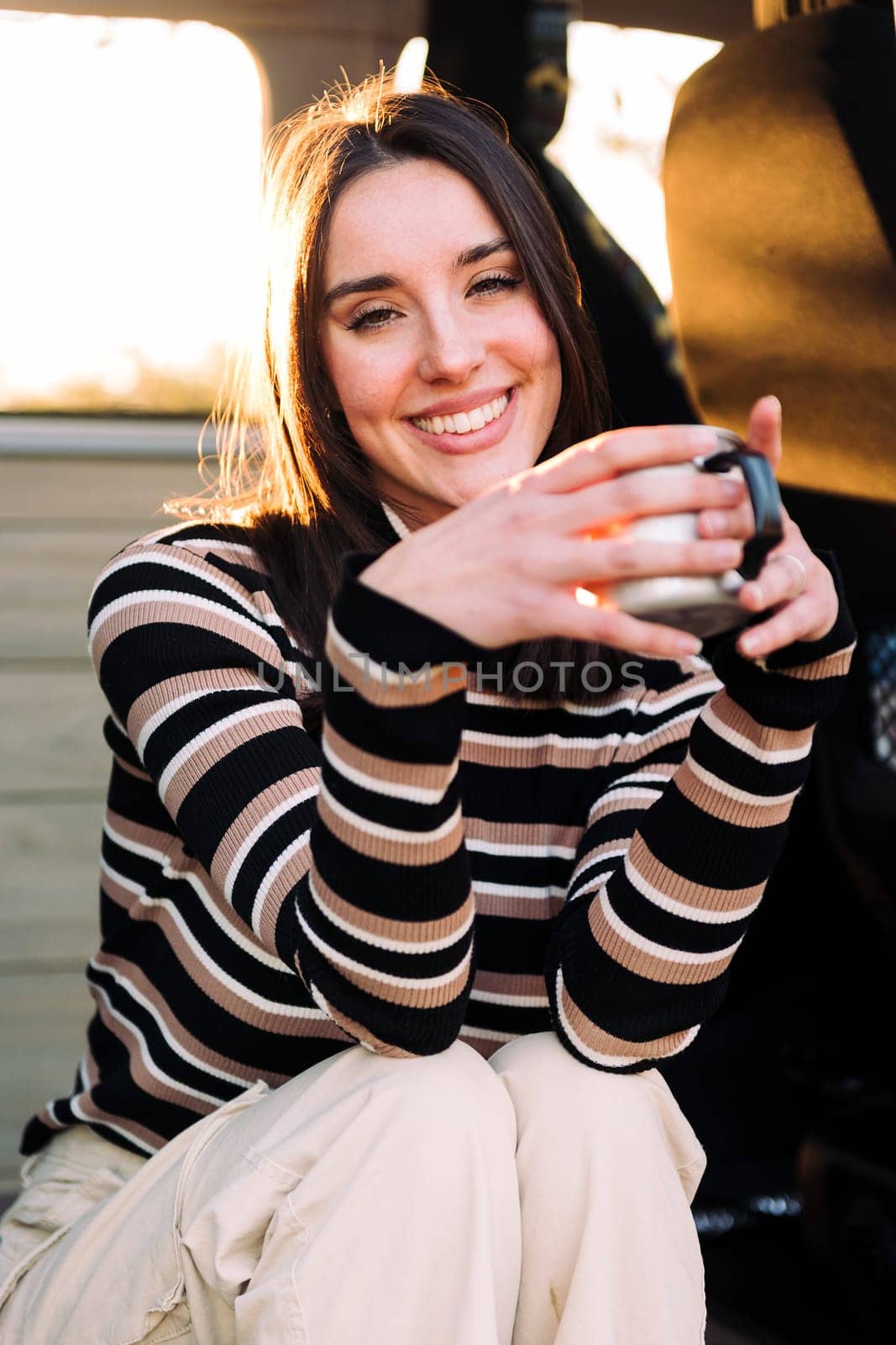 young caucasian woman smiling happy at sunset relaxing in a camper van with a cup of a hot drink her hand, concept of van life and weekend getaway