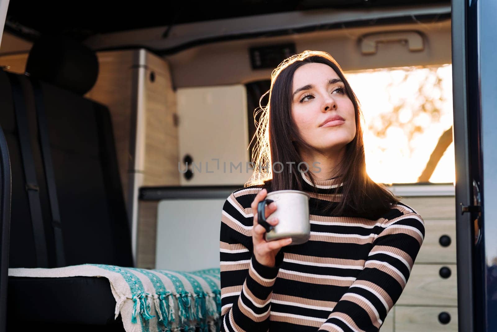 young caucasian woman chilling and enjoying a mug of a hot drink at sunset in a camper van, concept of van life and weekend getaway, copy space for text