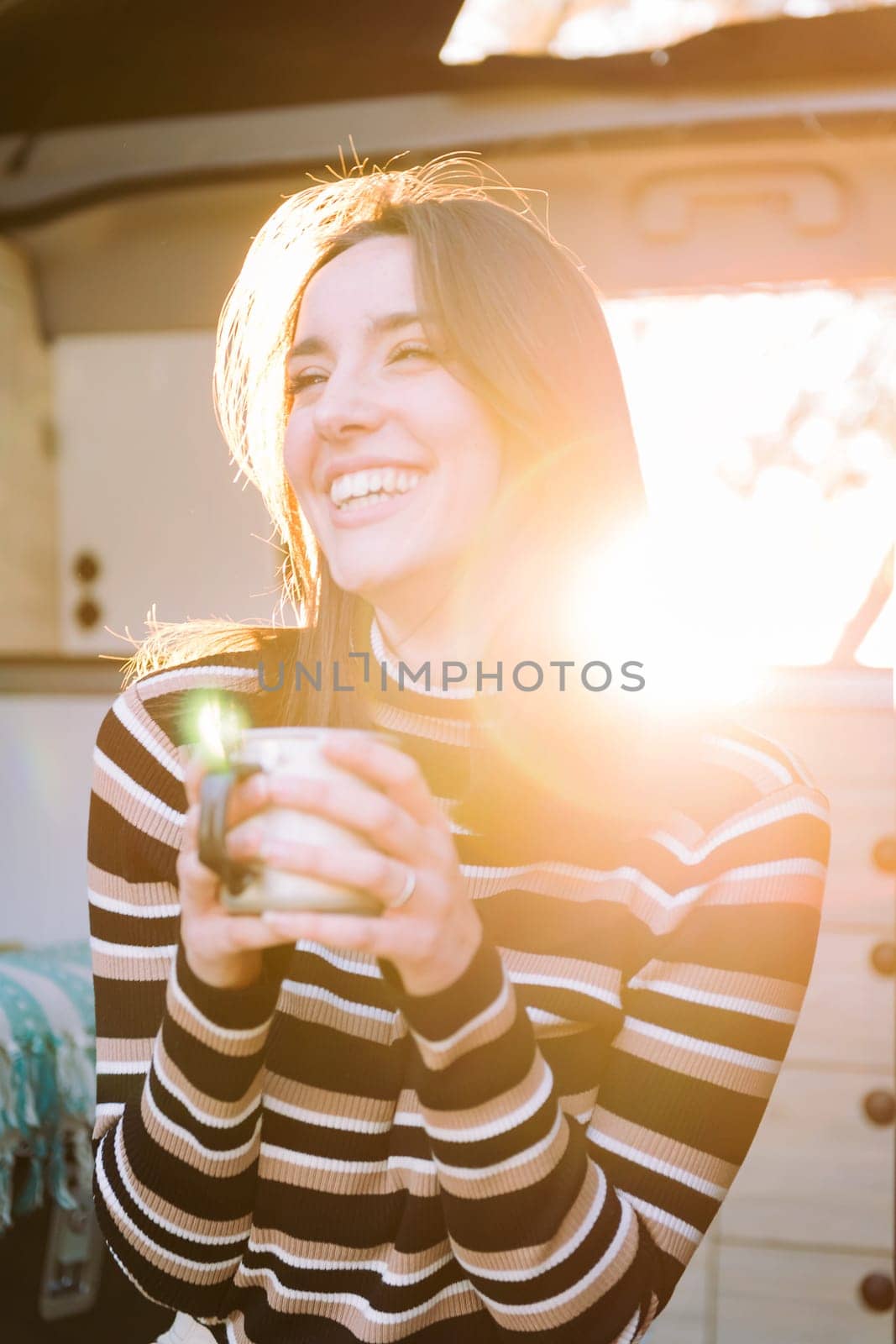 young woman laughing happy sitting in a camper van drinking a cup of coffee at sunset, concept of van life and weekend getaway