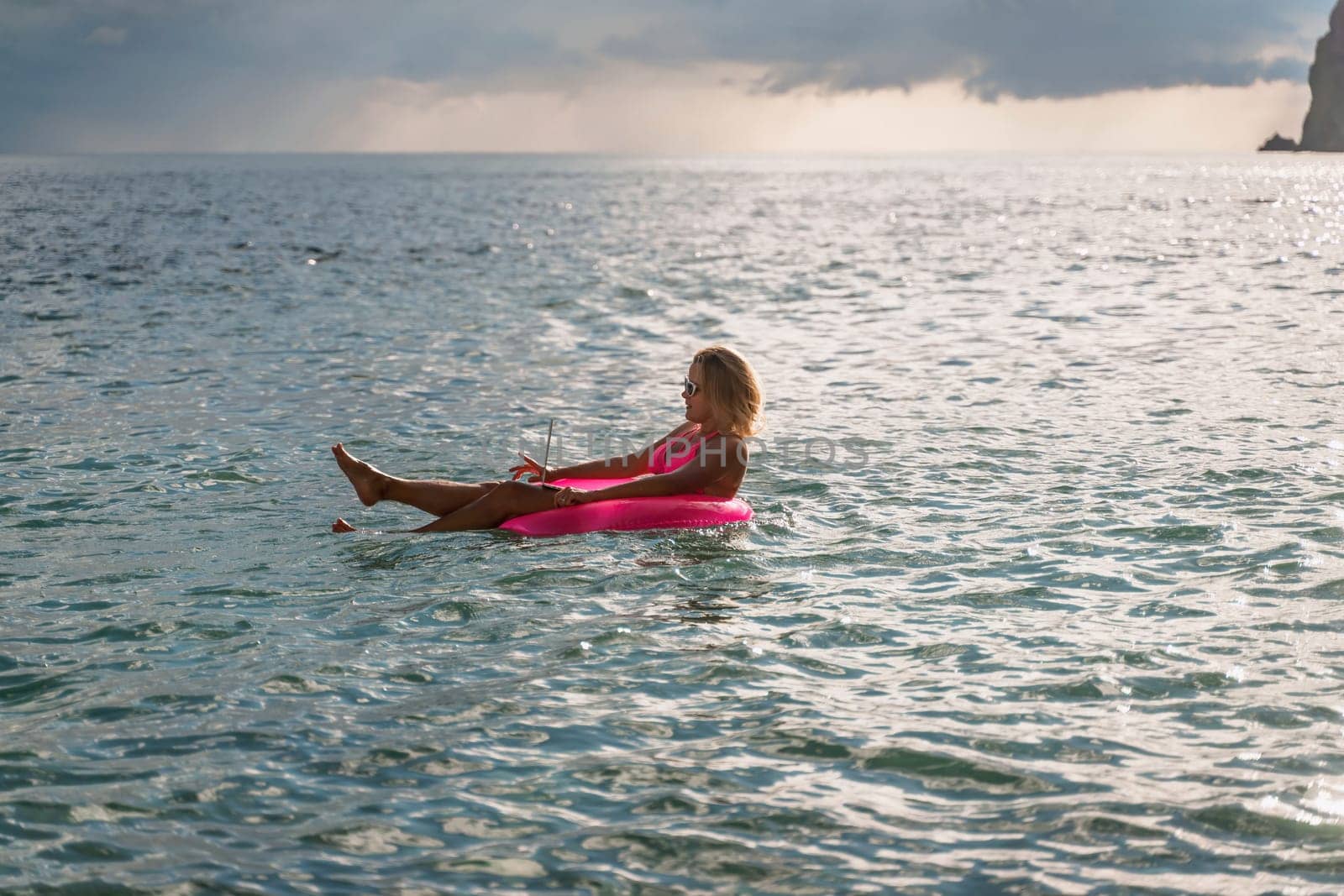 Woman works on laptop in sea. Freelancer, blond woman in sunglases floating on an inflatable big pink donut with a laptop in the sea at sunset. People summer vacation rest lifestyle concept