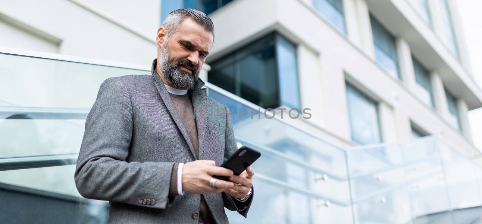 mature adult male financier with gray hair and a beard with a phone in his hands on the background of the building.