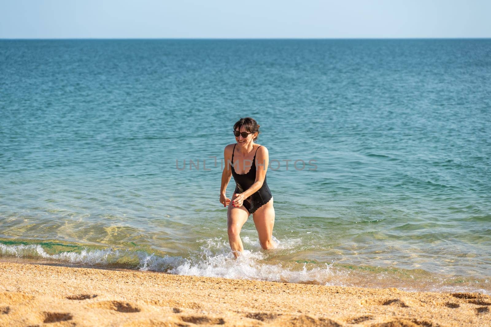 Woman sea swimsuit sand. The girl comes out of the sea in a black swimsuit after swimming. Alone on the beach on a sunny day