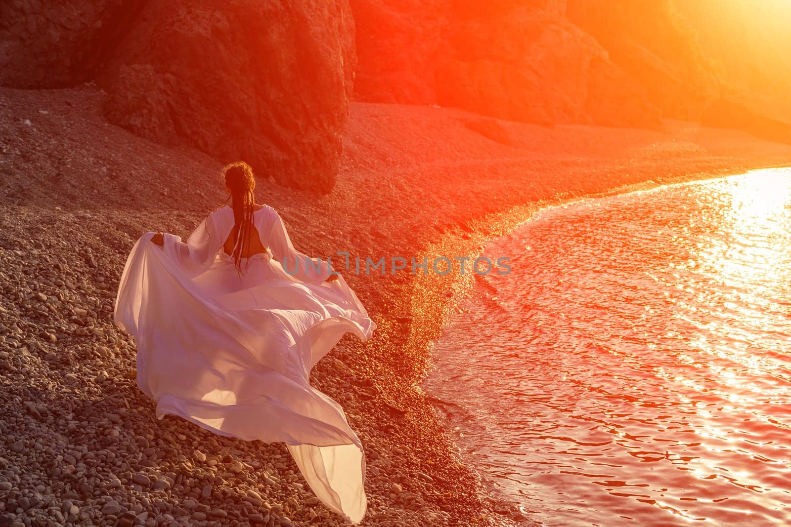 Mysterious woman silhouette long hair walks on the beach ocean water, sea nymph wind listens to the wave. Throws up a long white dress, a divine sunset. Artistic photo from the back without a face.