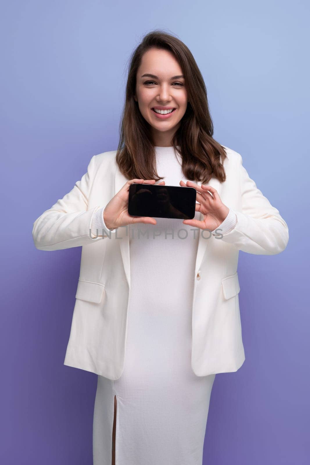 positive energetic young brunette with long hair woman in white dress holding smartphone with mockup.