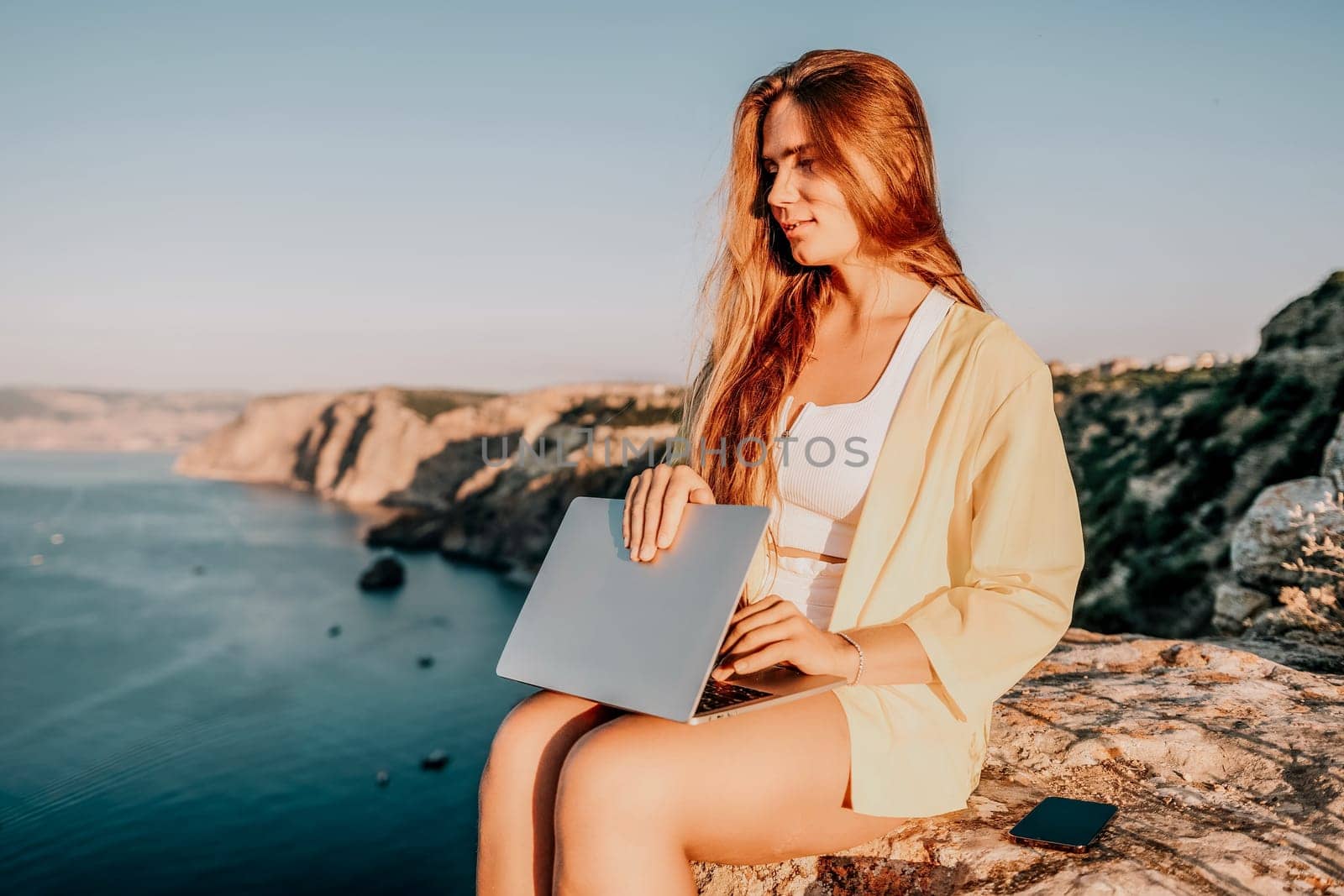 Successful business woman in yellow hat working on laptop by the sea. Pretty lady typing on computer at summer day outdoors. Freelance, travel and holidays concept.