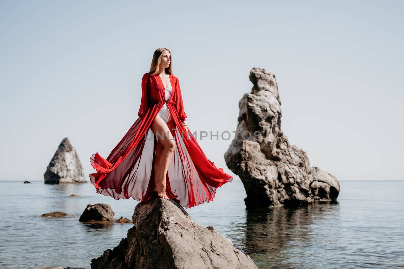 Woman travel sea. Young Happy woman in a long red dress posing on a beach near the sea on background of volcanic rocks, like in Iceland, sharing travel adventure journey by panophotograph