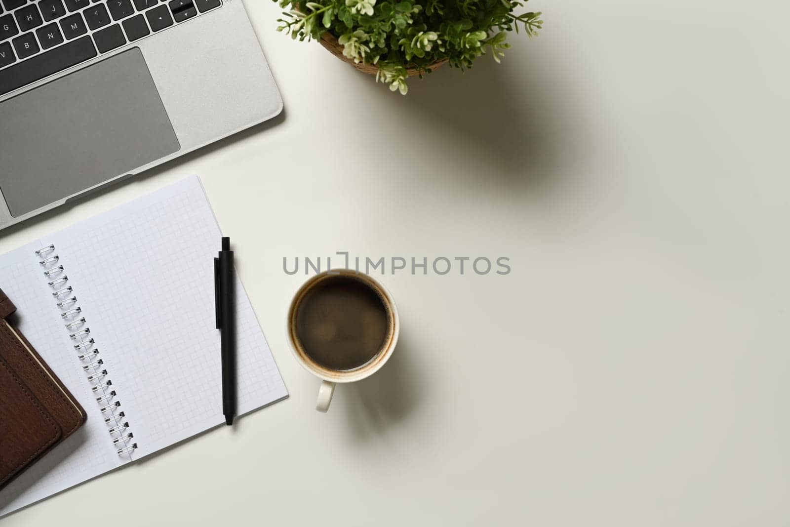 A cup of coffee, laptop, blank notepad and potted plant on white office desk. Top view with coy space.