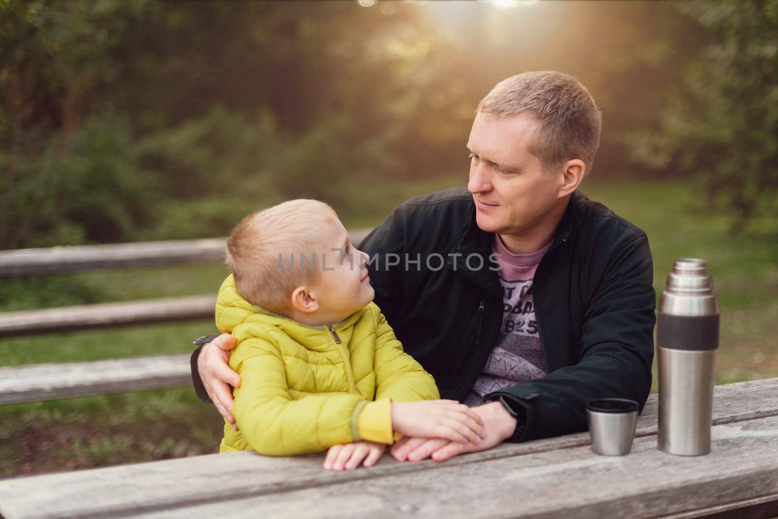 Happy family: father and child boy son playing and laughing in autumn park, sitting on wooden bench. Father and little kid having fun outdoors, playing together. Father and son sitting on a bench and talking. dad son park bench table autumn thermos.
