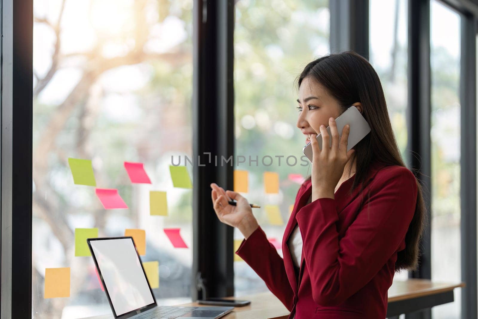 businesswoman talking on cellphone and work with laptop, writing notes on colorful sticky papers in office, consulting client by phone call.