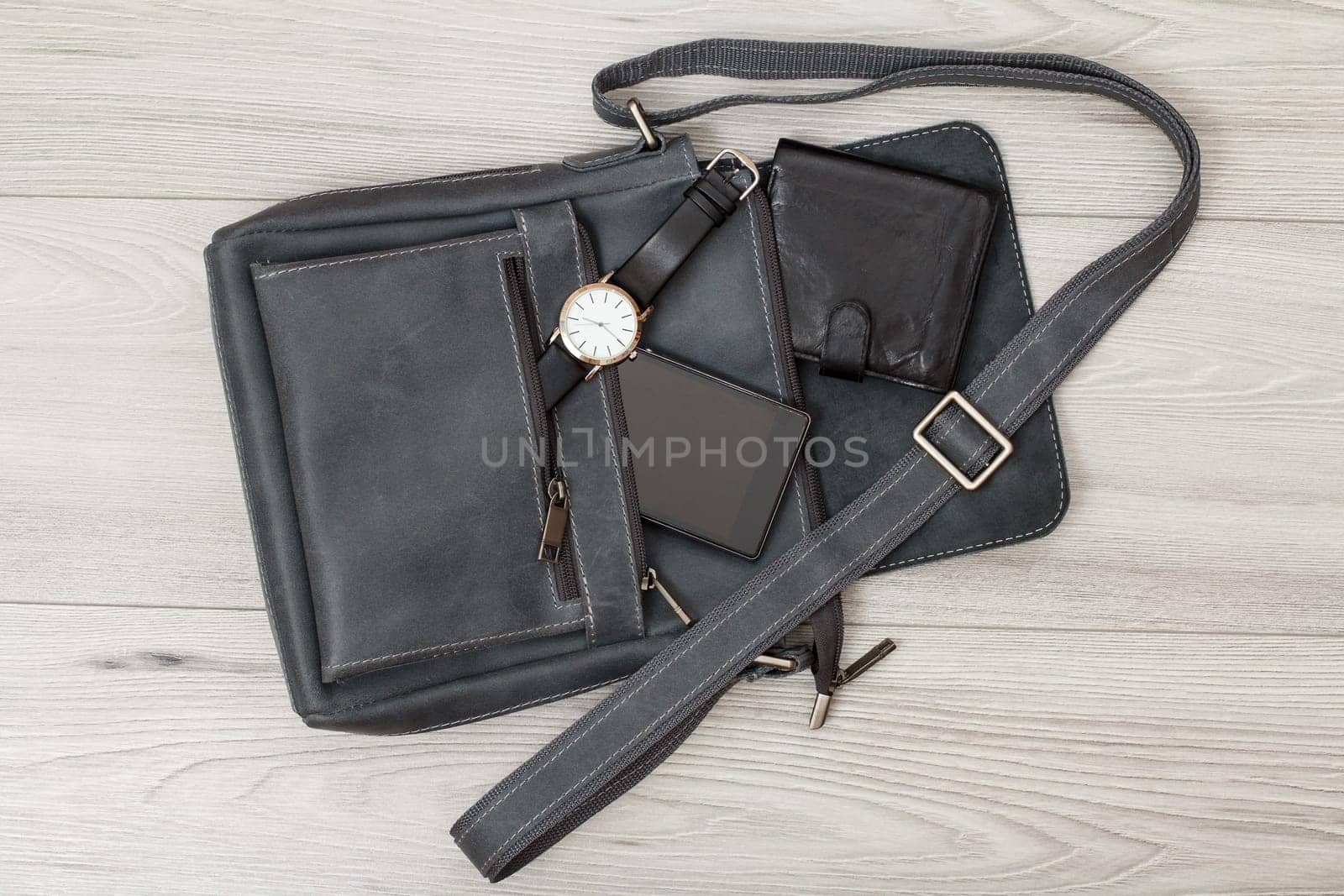 Leather shoulder bag for men with mobile phone, watch and wallet on it with gray wooden background. Men's accessories. Top view.