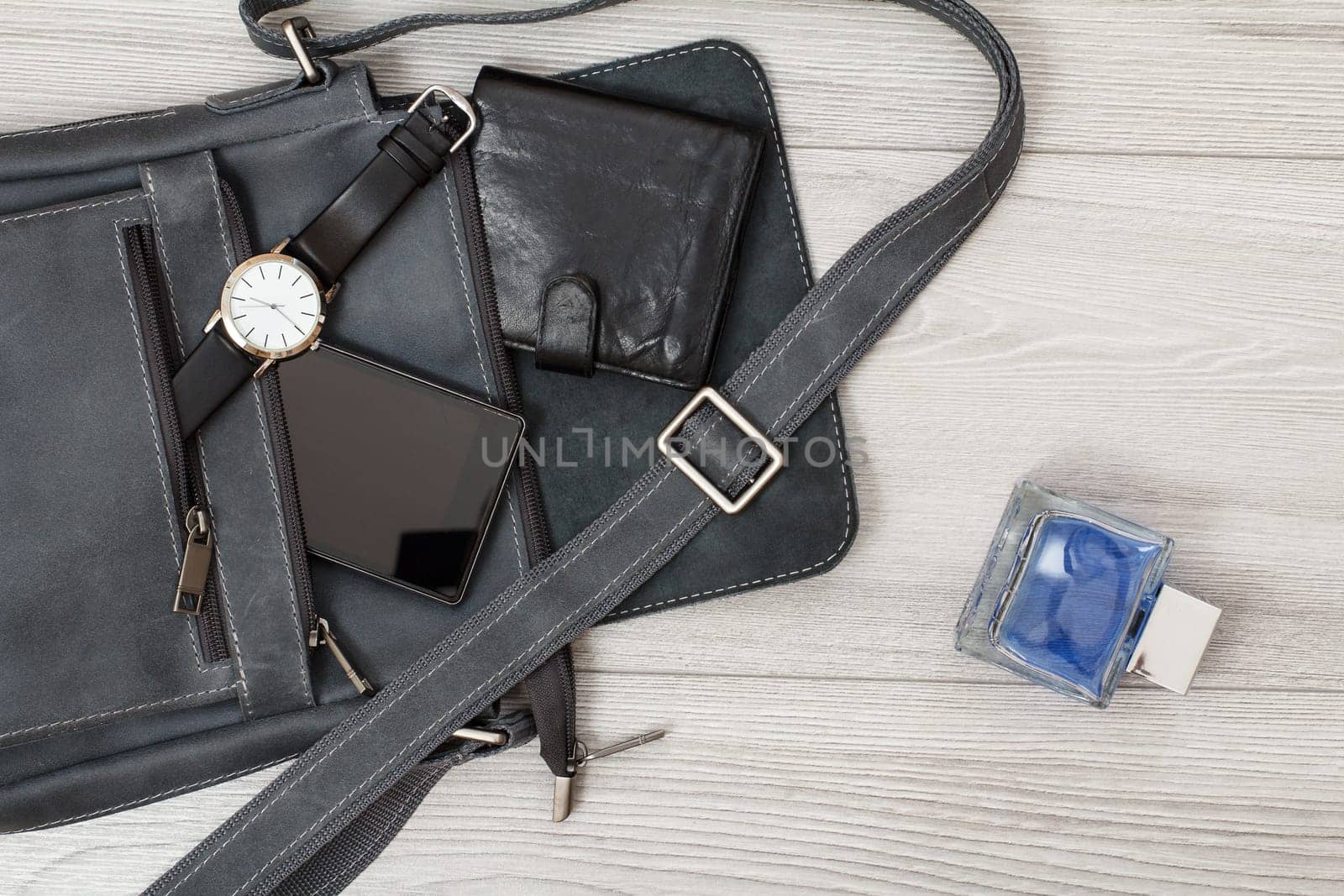 Leather shoulder bag for men with mobile phone, watch and wallet on it, men's cologne on gray wooden background. Men's accessories. Top view.