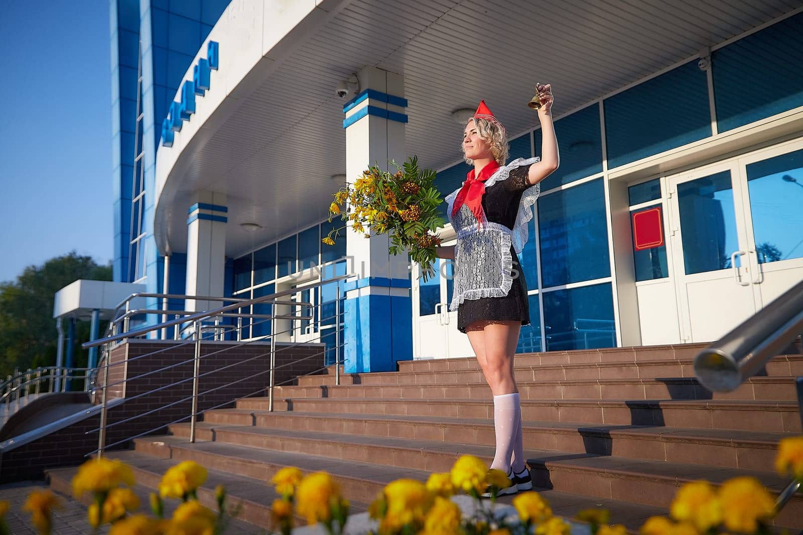 A Girl in black uniform, white apron, red tie on steps of school with bouquet of flowers and bell. Nostalgia photo shoot of teenager of female pioneer from USSR costume for September 1 or graduation
