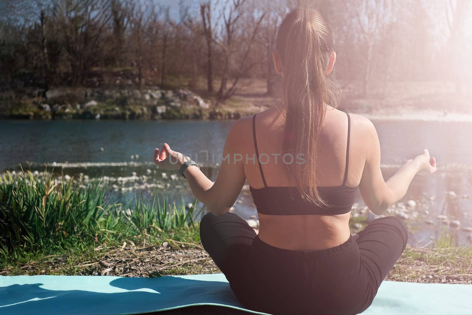Young woman does yoga on lawn in park near the river, stretching on fitness mat, back view. Copy space