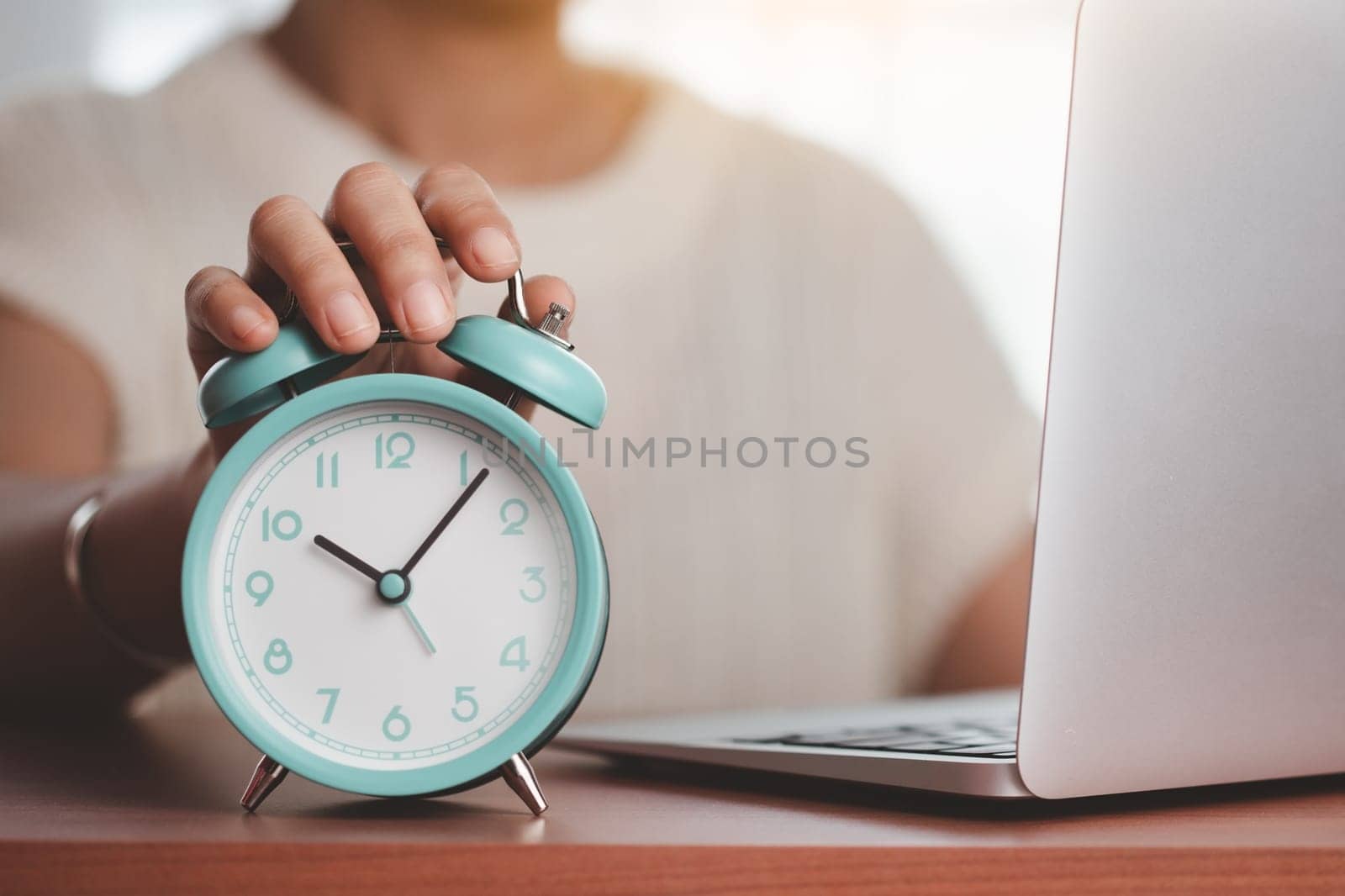 Asian woman's hand reaching for an alarm clock to check the time on desk with a computer laptop for the concept of work, study and time management.