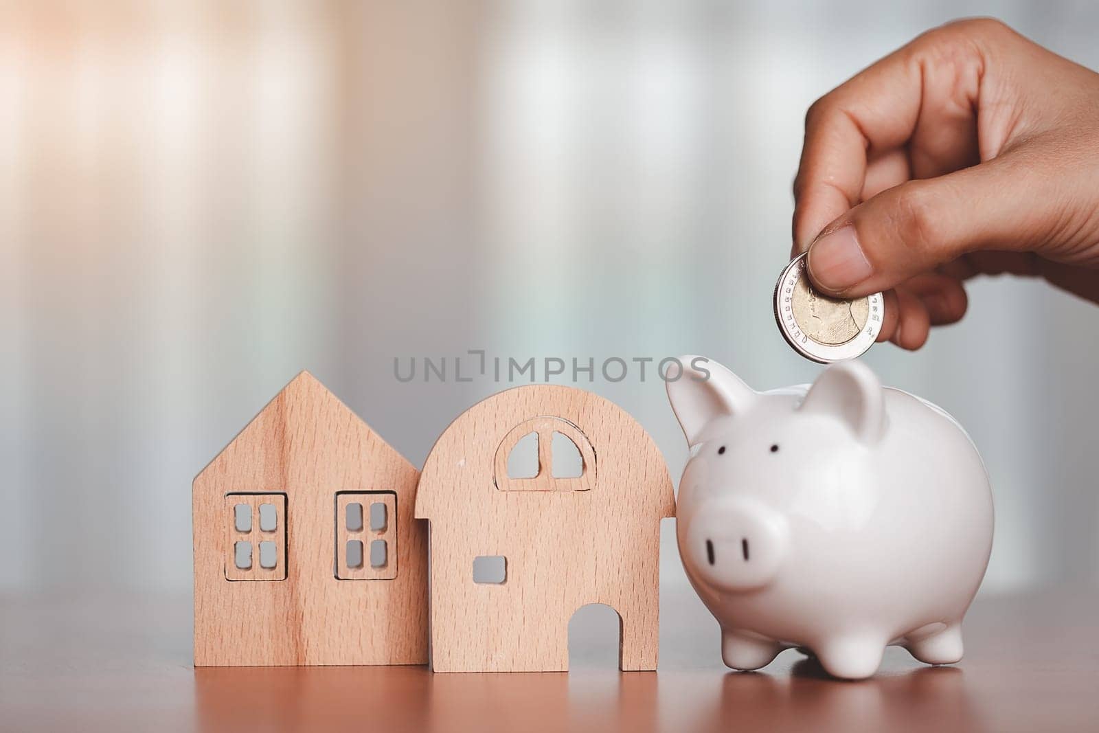 Asian women's hand saving a coin into piggy bank with wooden house model on the table for business, finance, saving money and property investment concept.