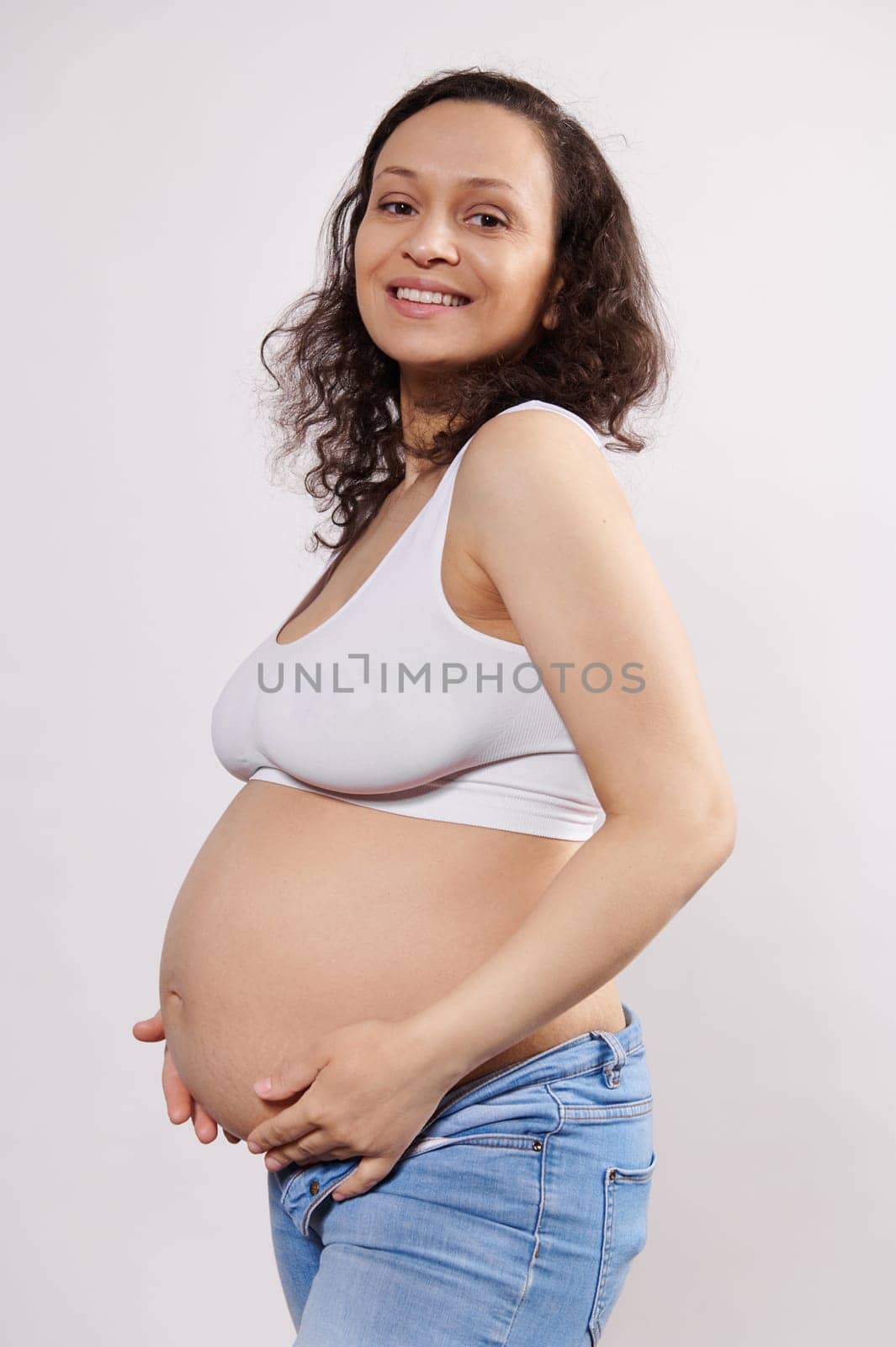 Attractive happy pregnant ethnic woman holding hands on her tummy, smiling looking at camera, isolated white background. by artgf
