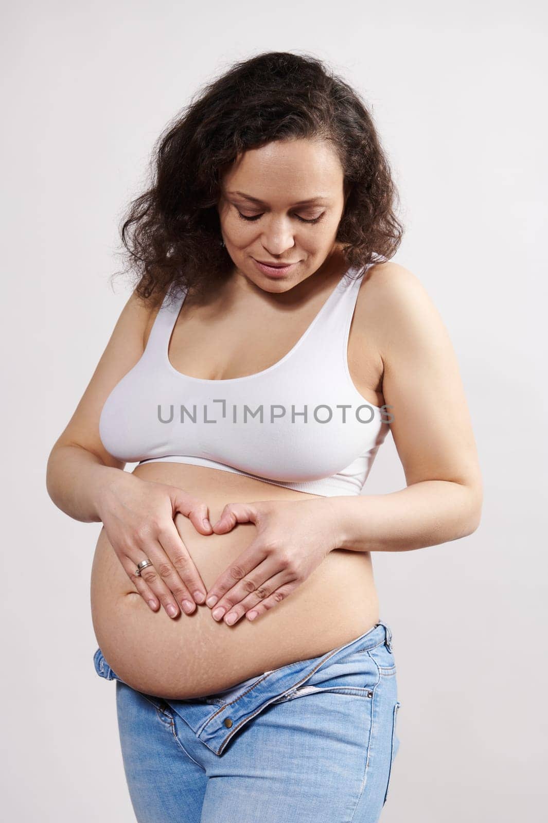 Pregnant woman in white underwear and denim jeans, putting her hands on her belly, making heart shape, isolated on white by artgf