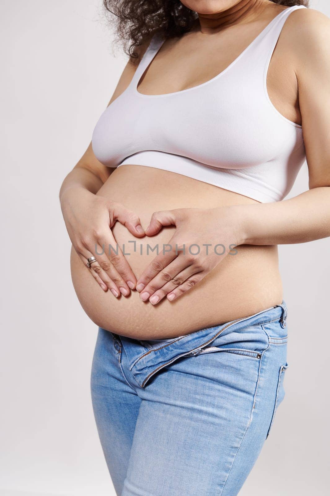 Closeup pregnant woman putting her hands on her naked belly, making heart shape from fingers, isolated on white background. Delightful gravid female expecting a baby. Happy carefree pregnancy 30 week