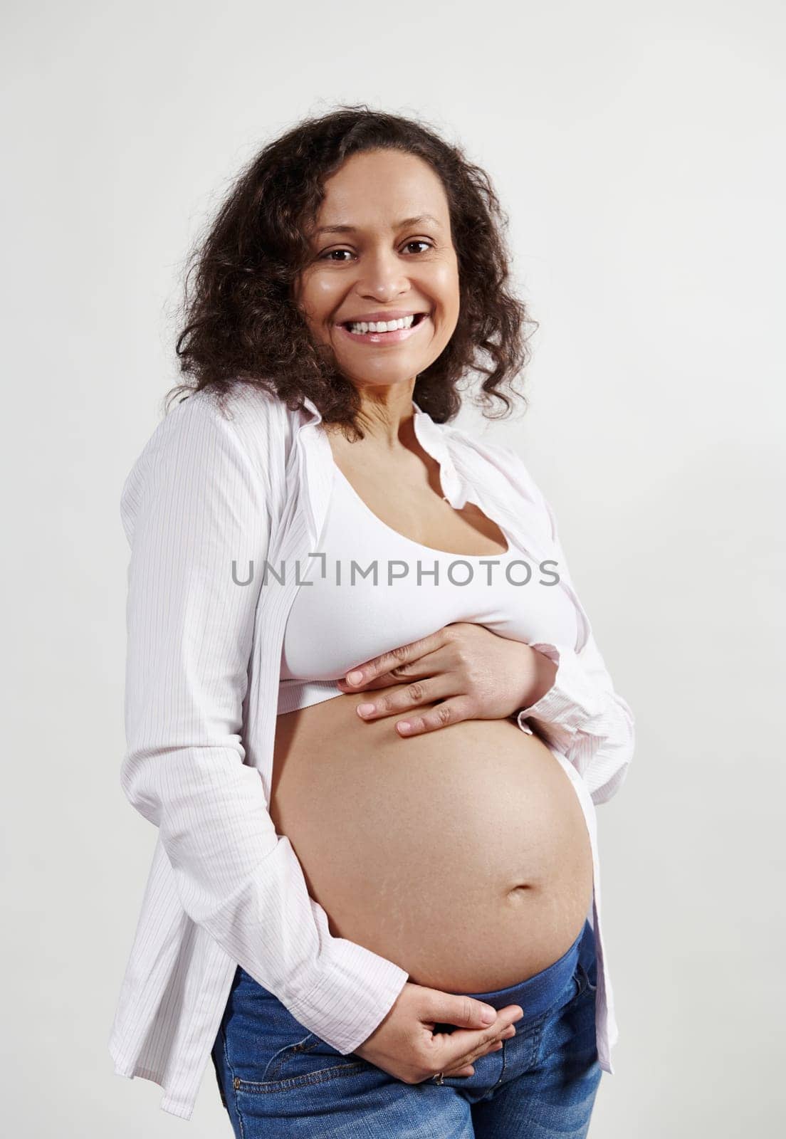 African American pregnant pretty woman in denim jeans and white shirt, smiling looking at camera, expressing positive emotions, caressing her bare belly, feeling first baby kicks, isolated background