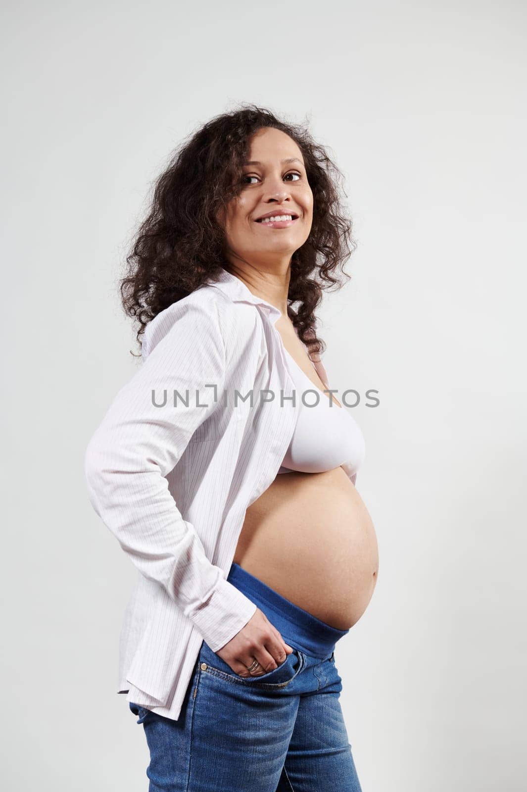 Sexy curly haired pregnant woman in sixth month of pregnancy , smiling, posing with bare belly isolated white background by artgf