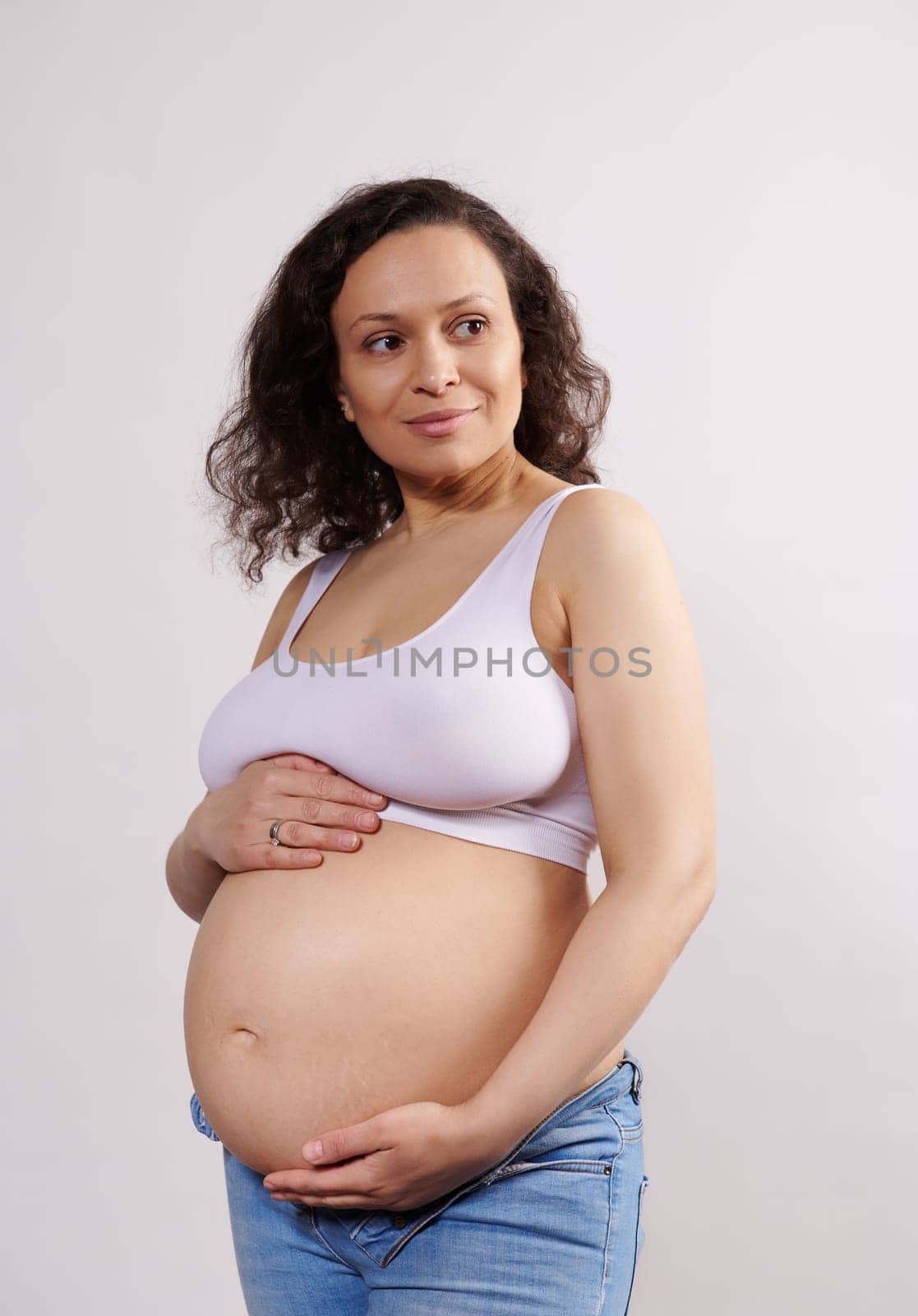 Confident delightful Latin American gravid young woman 35-39 years old, smiling cutely looking aside, caressing her pregnant belly, enjoying happy and carefree pregnancy, isolated on white background