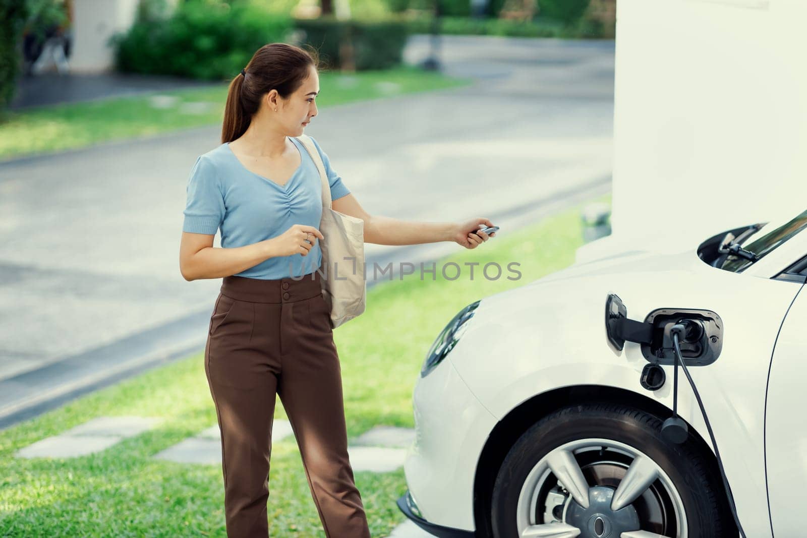 Progressive woman install cable plug to her electric car with home charging station. Concept of the use of electric vehicles in a progressive lifestyle contributes to clean environment.