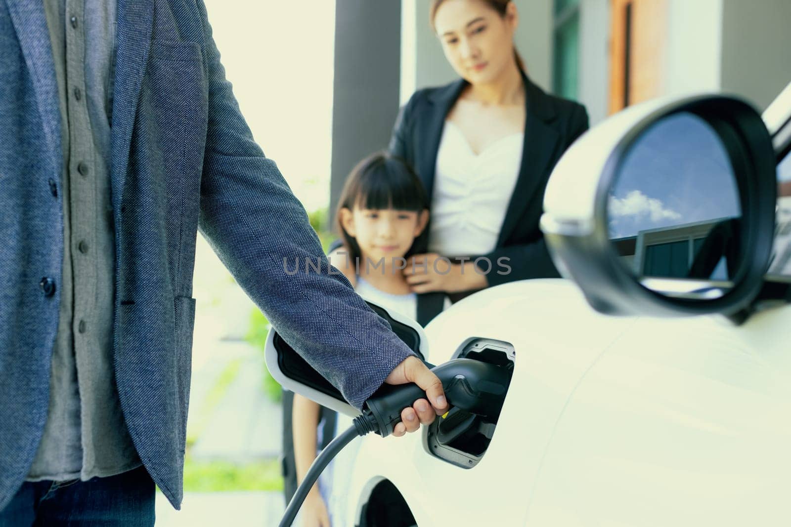 Focus closeup hand charging electric car, insert charger device into electric vehicle as progressive lifestyle concept of alternative green energy technology with blurred family in the background.