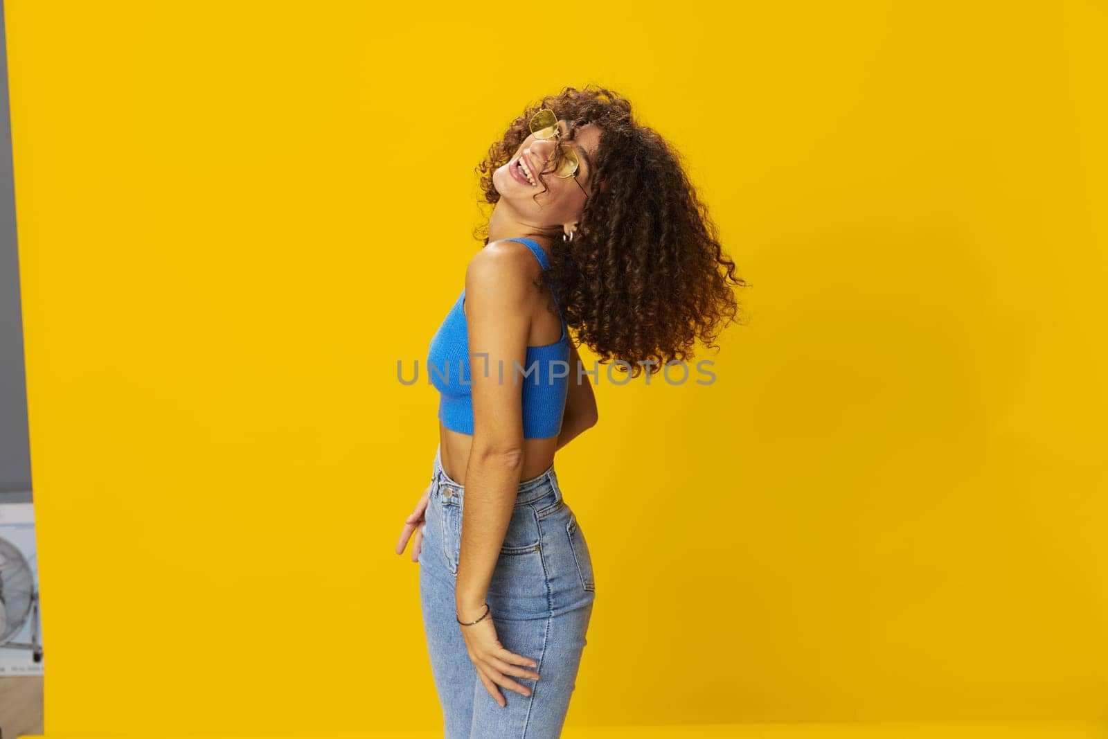 Woman with curly afro hair in a blue T-shirt on a yellow background dancing flying hair with sunglasses yellow, hand signs, look into the camera, smile with teeth and happiness, copy space. High quality photo