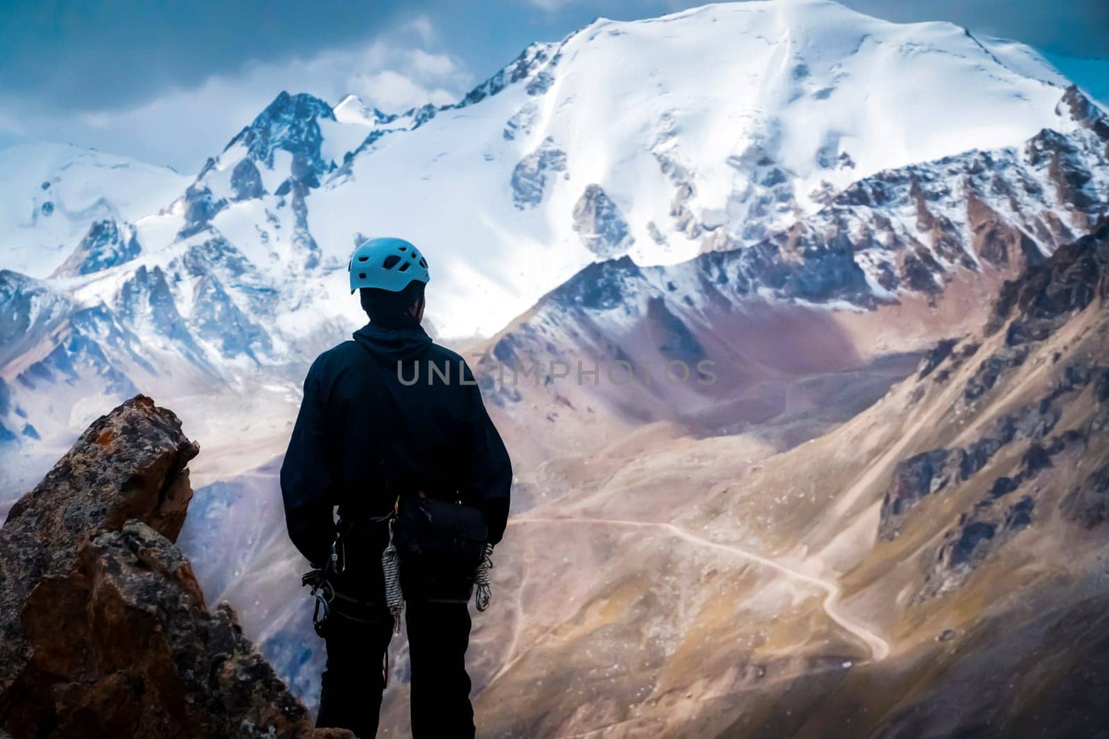 A young man traveler is engaged in mountaineering. Hiker in a helmet, with a rope climbs to the top, against the backdrop of a stunning view with snow-capped mountains.