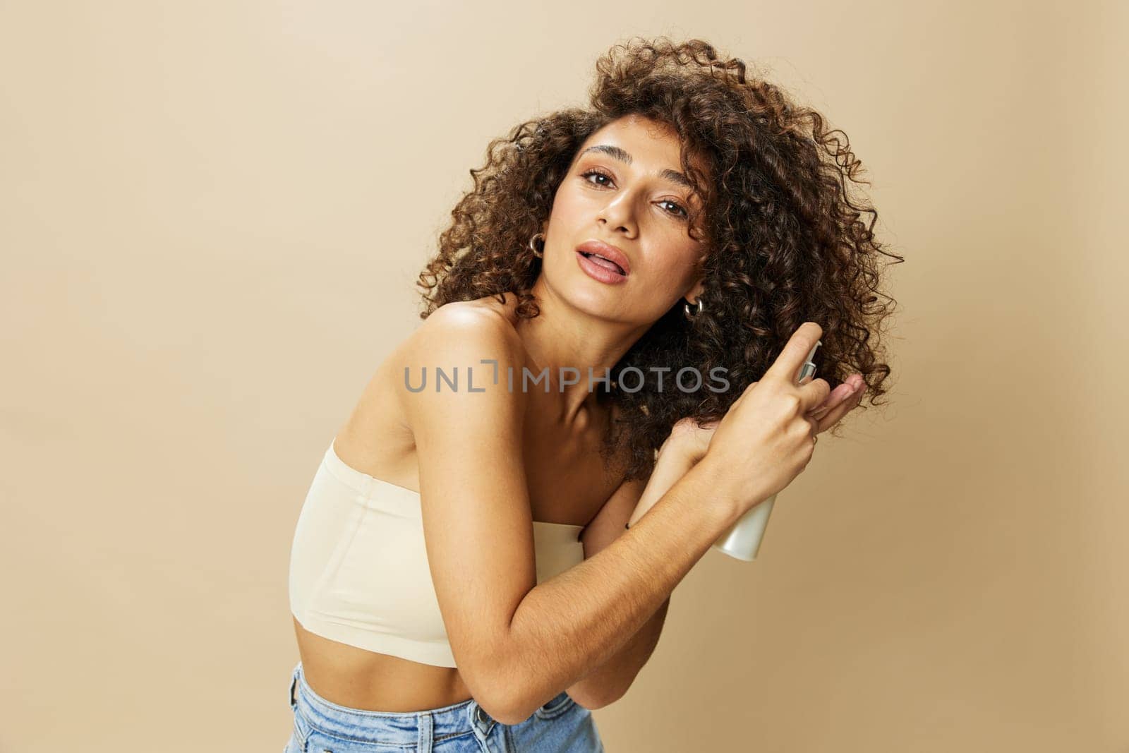 Woman applies cream and balm to her curly hair, the concept of protection and care with salon products, a healthy look, a smile with teeth on a beige background. High quality photo