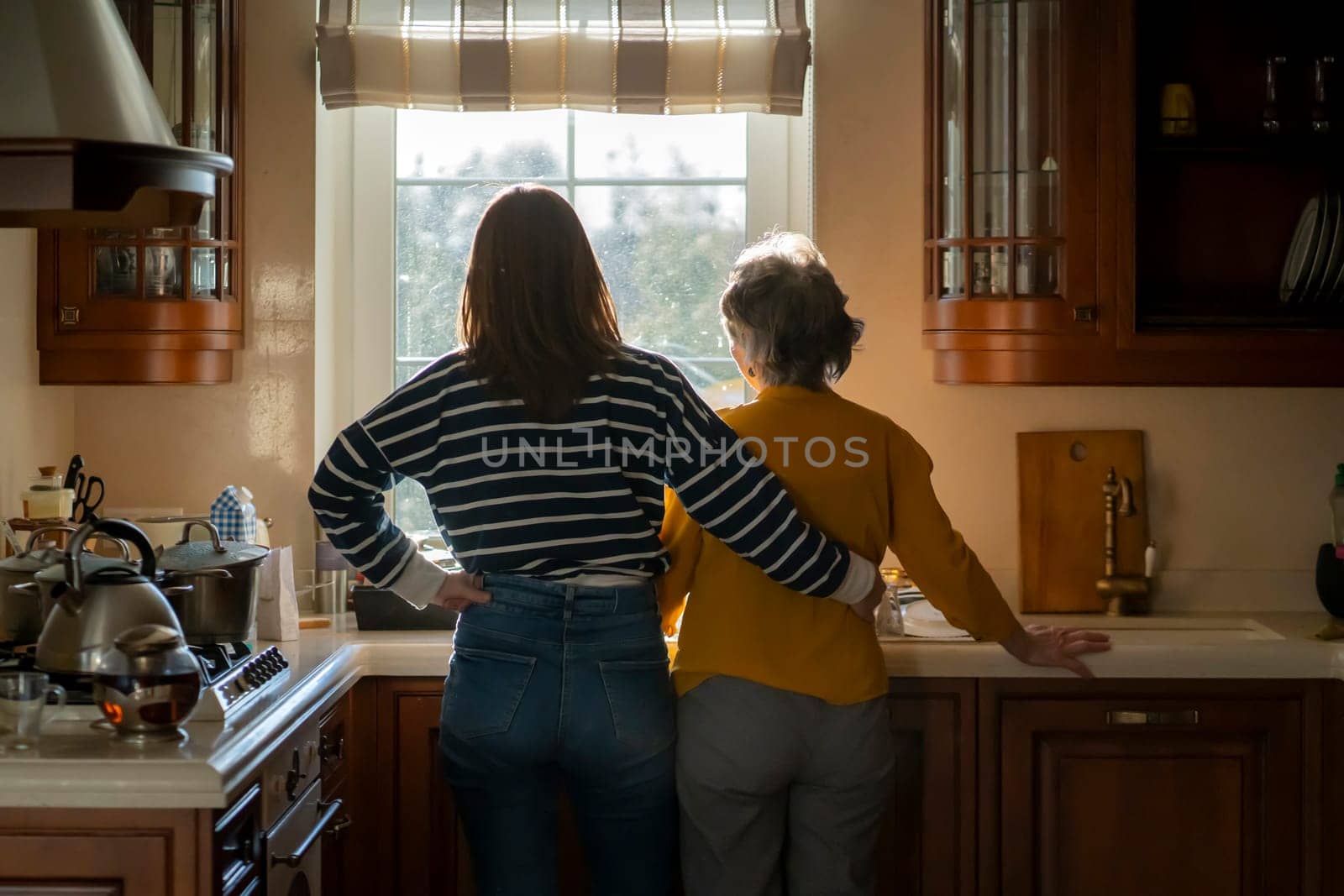 The daughter hugs her elderly mother, the granddaughter and her grandmother enjoy life, look out the window at a beautiful view from their cozy home, spend time preparing together in the kitchen.