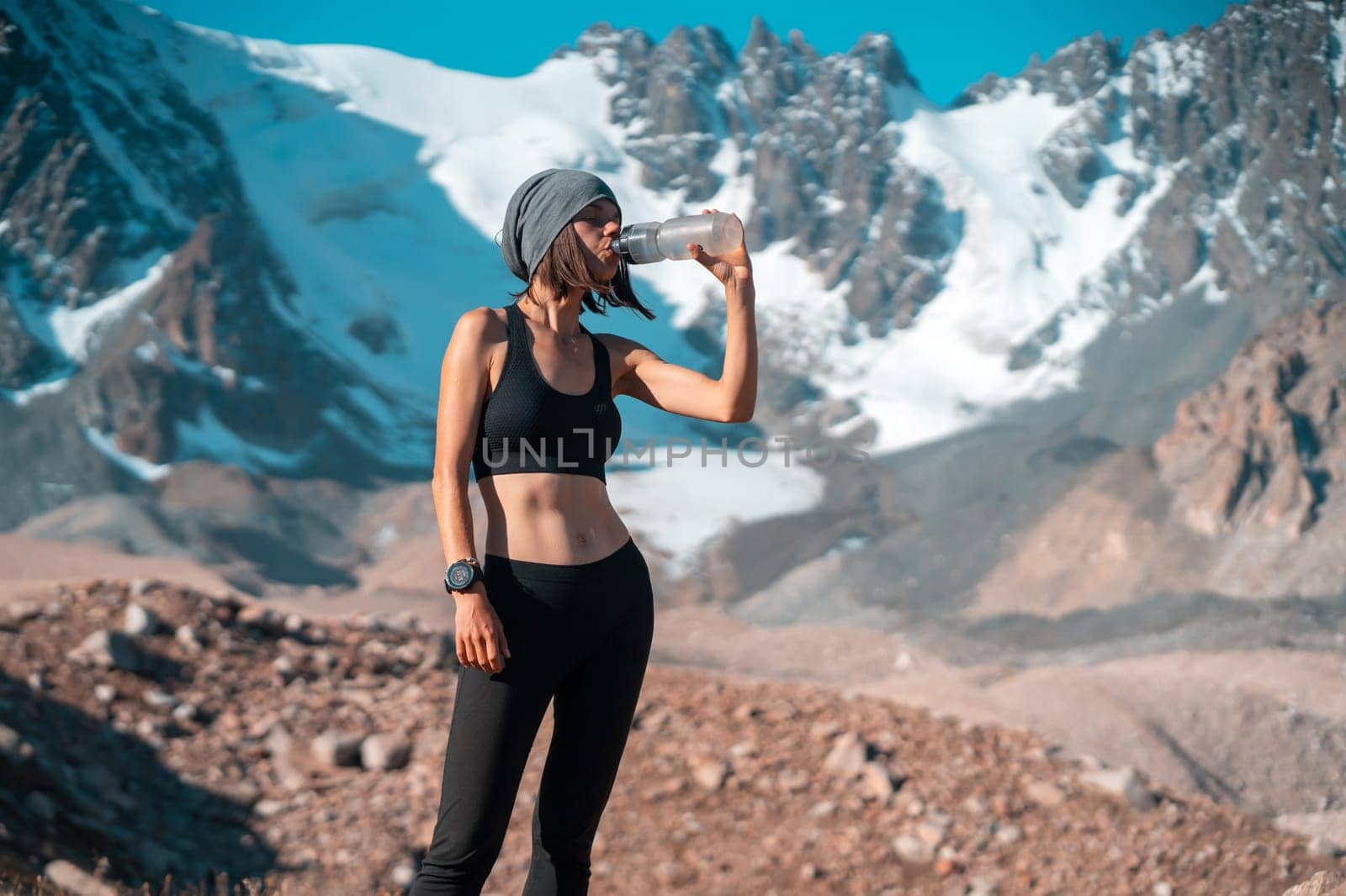 A young girl of athletic build drinks water, isotonic from a bottle during training on trail running in the snow-capped mountains. Runner is making workout outdoor.