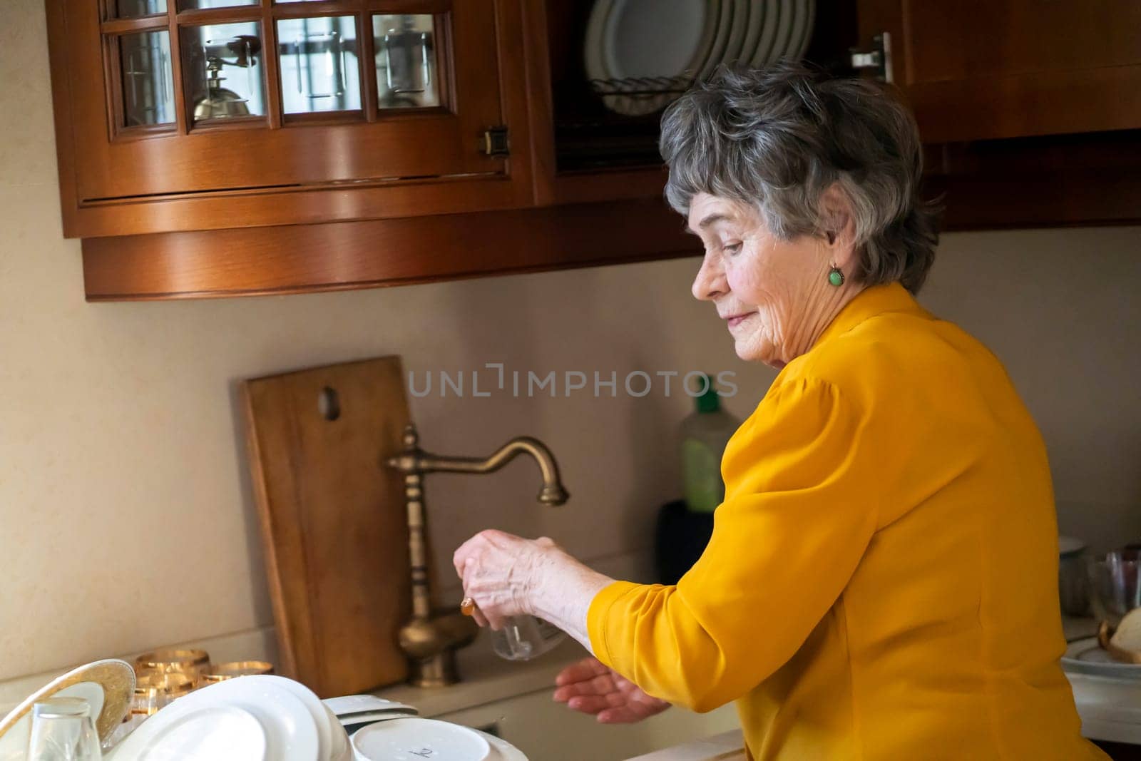 Elderly retired gray-haired woman having a good time in her house, active in her old 80 age, washing dishes in a cozy kitchen, cleaning. A happy grandmother in a yellow blouse, young at heart.