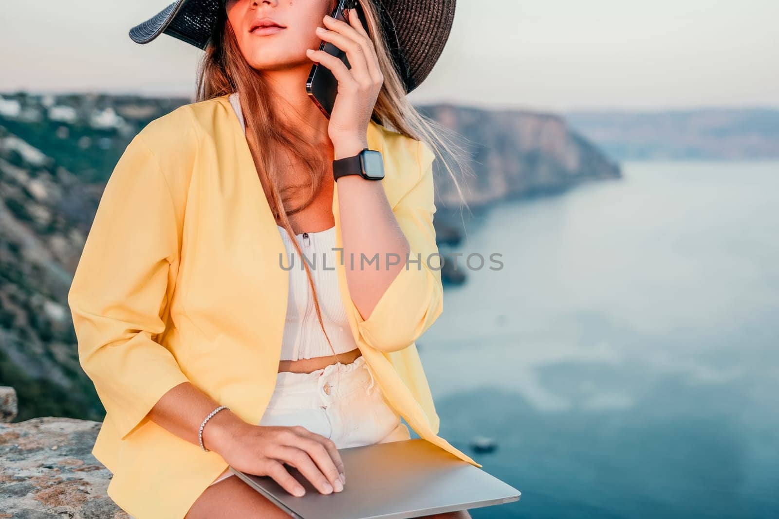 Successful business woman in yellow hat working on laptop by the sea. Pretty lady typing on computer at summer day outdoors. Freelance, travel and holidays concept.