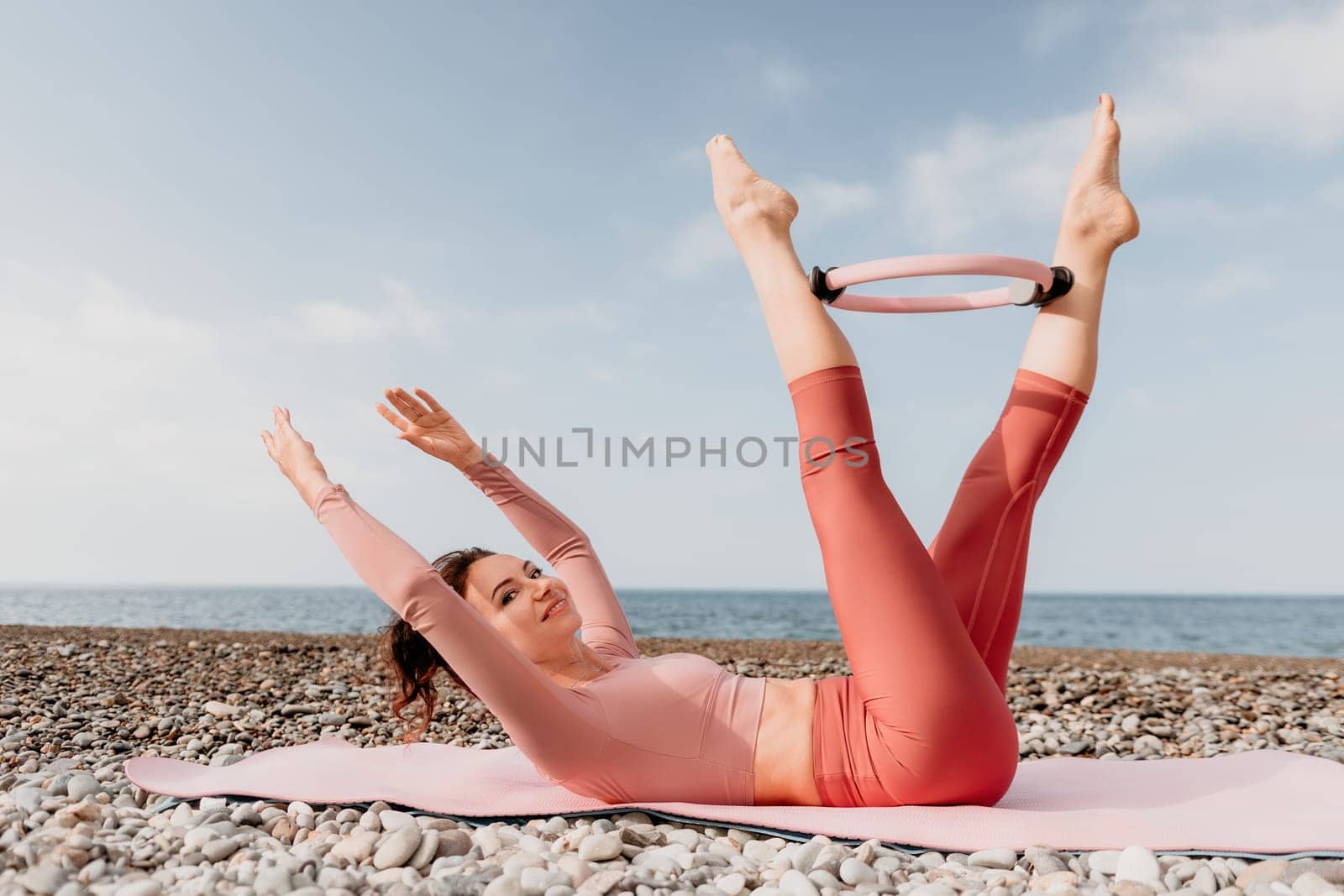 Woman sea pilates. Sporty happy middle aged woman practicing fitness on beach near sea, smiling active female training with ring on yoga mat outside, enjoying healthy lifestyle, harmony and meditation by panophotograph