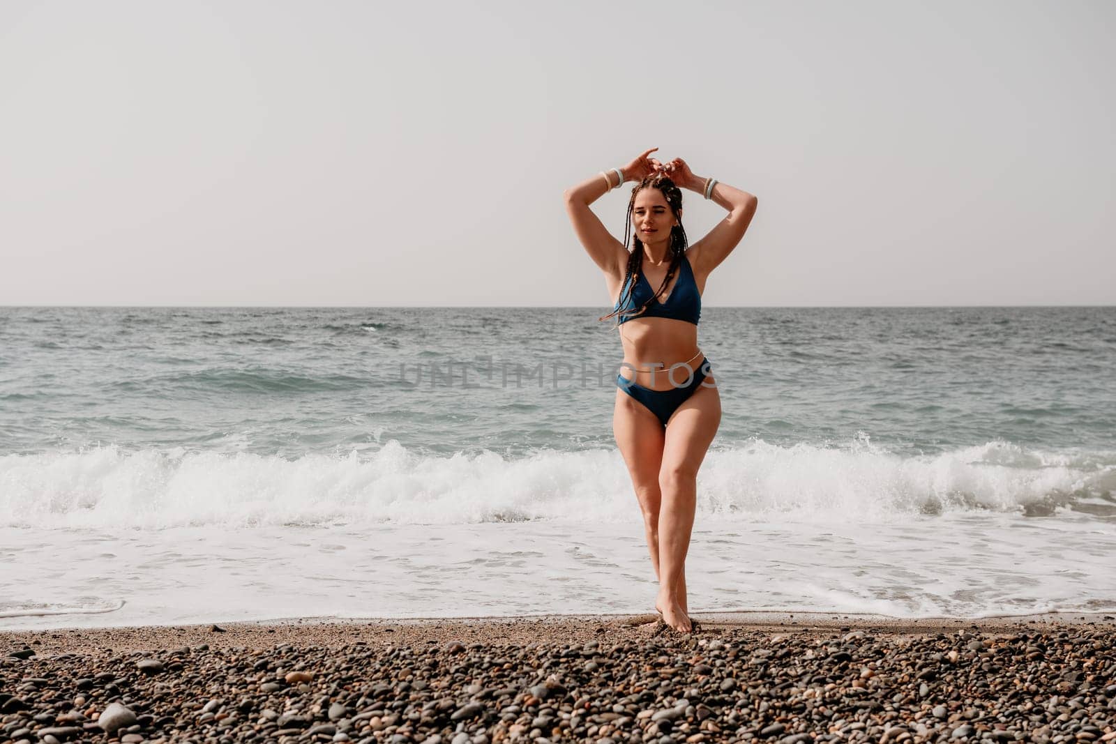 Beach vacation. Hot beautiful woman in sunhat and bikini standing with her arms raised to her head enjoying looking view of beach ocean on hot summer day.