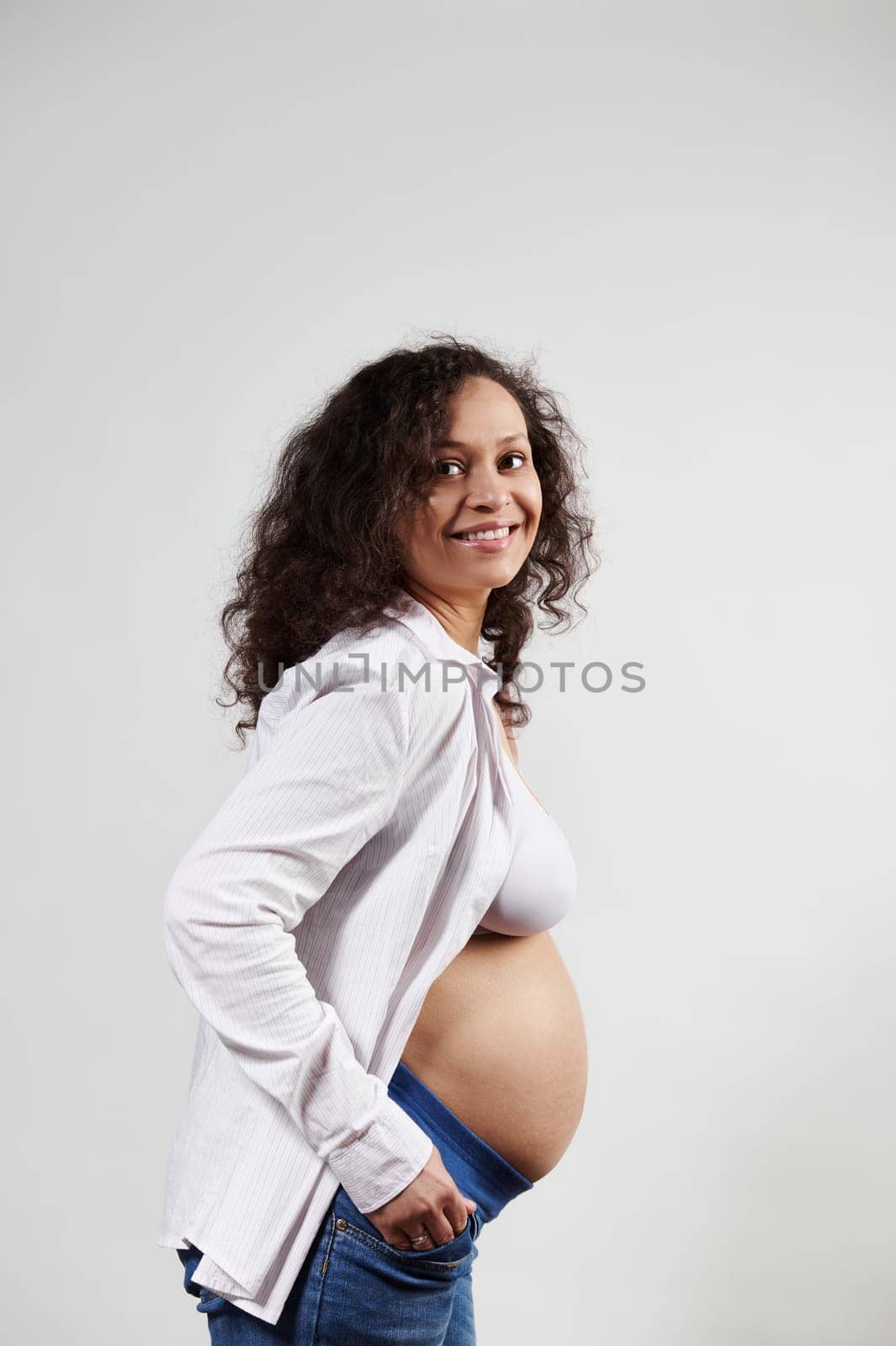 Curly happy pregnant woman 30s, smiling posing with hands in jeans pockets and bare belly, on isolated white background by artgf