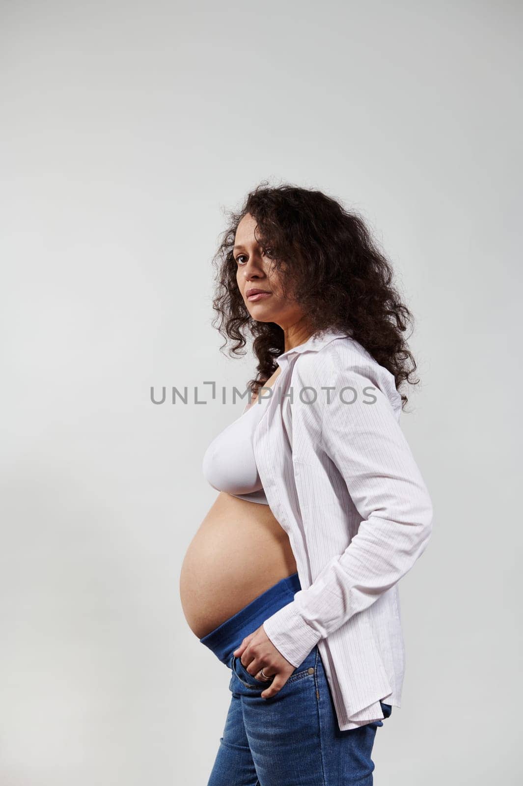 Vertical studio portrait of curly haired brunette pregnant woman, wearing blue jeans and unbuttoned white shirt, posing bare belly over isolated white background. Happy pregnancy and maternity concept