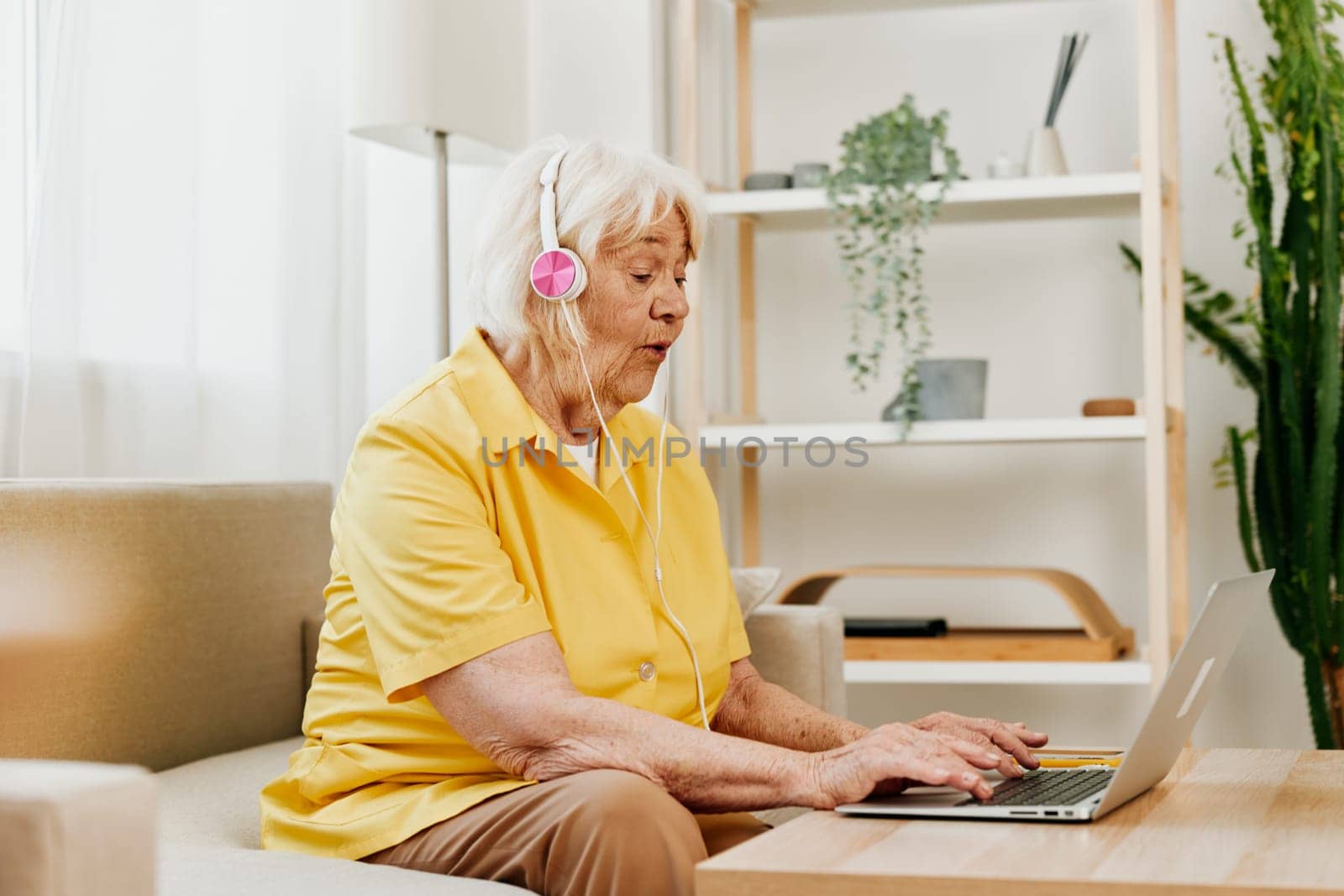 An elderly woman with headphones and a laptop sits on the couch at home and works. by SHOTPRIME