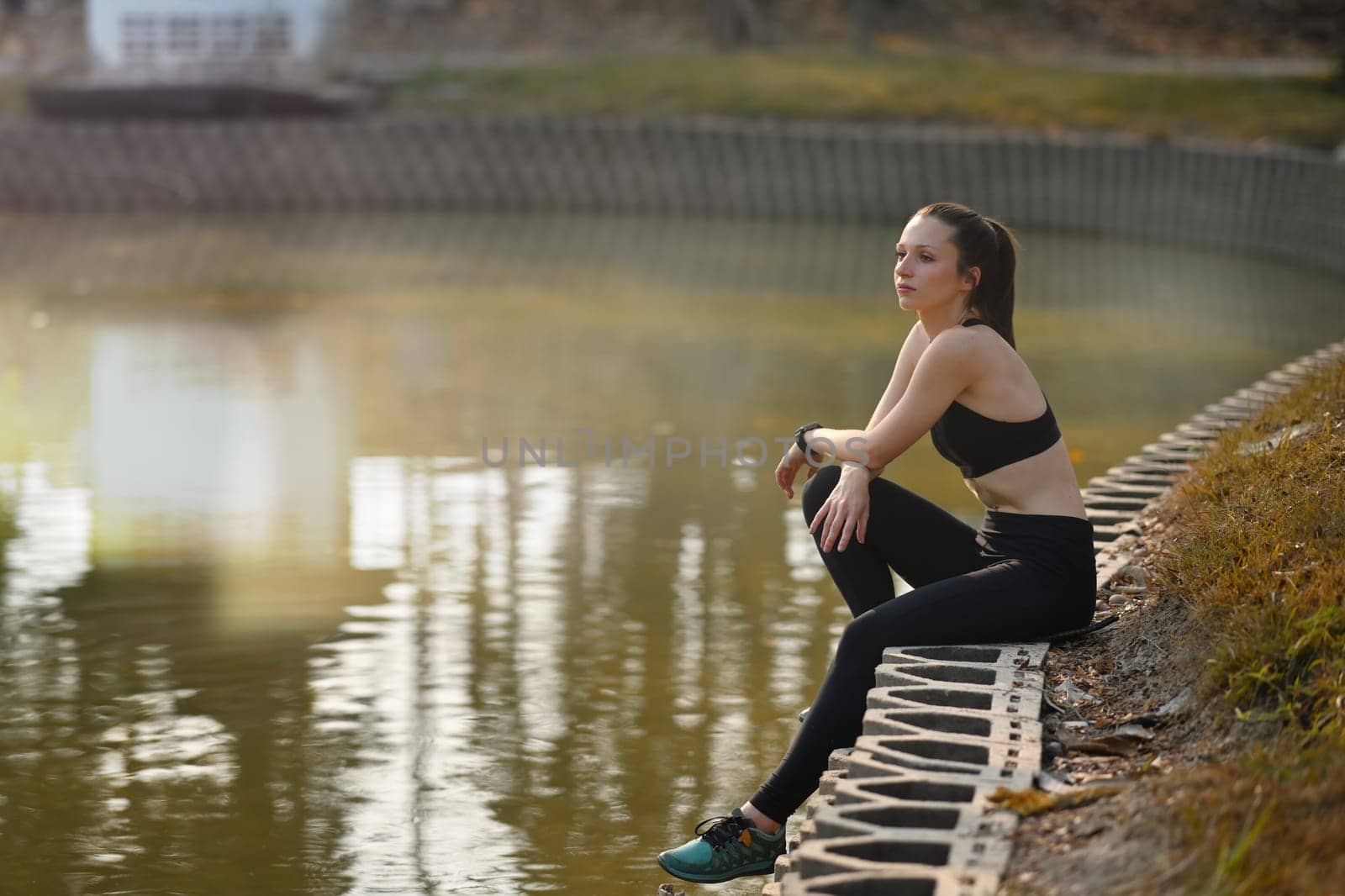 Fitness young caucasian woman sitting near river, resting after morning workout outdoor at summer park by prathanchorruangsak