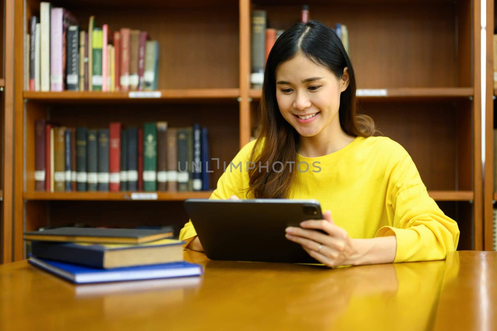 Smiling female college students preparing for exam, searching information on digital tablet in library.