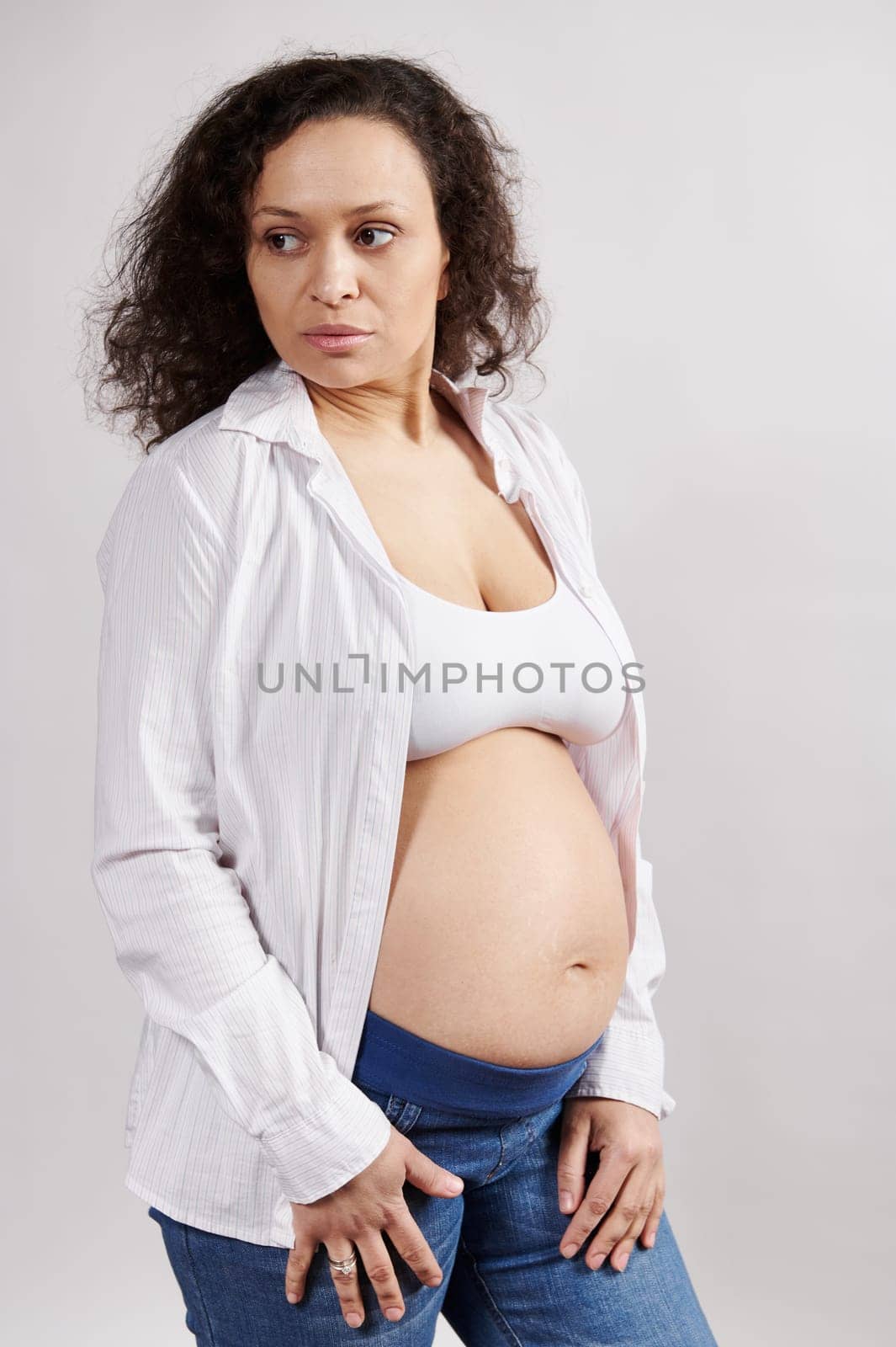Confident serene pregnant woman, wearing unbuttoned white shirt and blue denim, looking aside, isolated white background by artgf