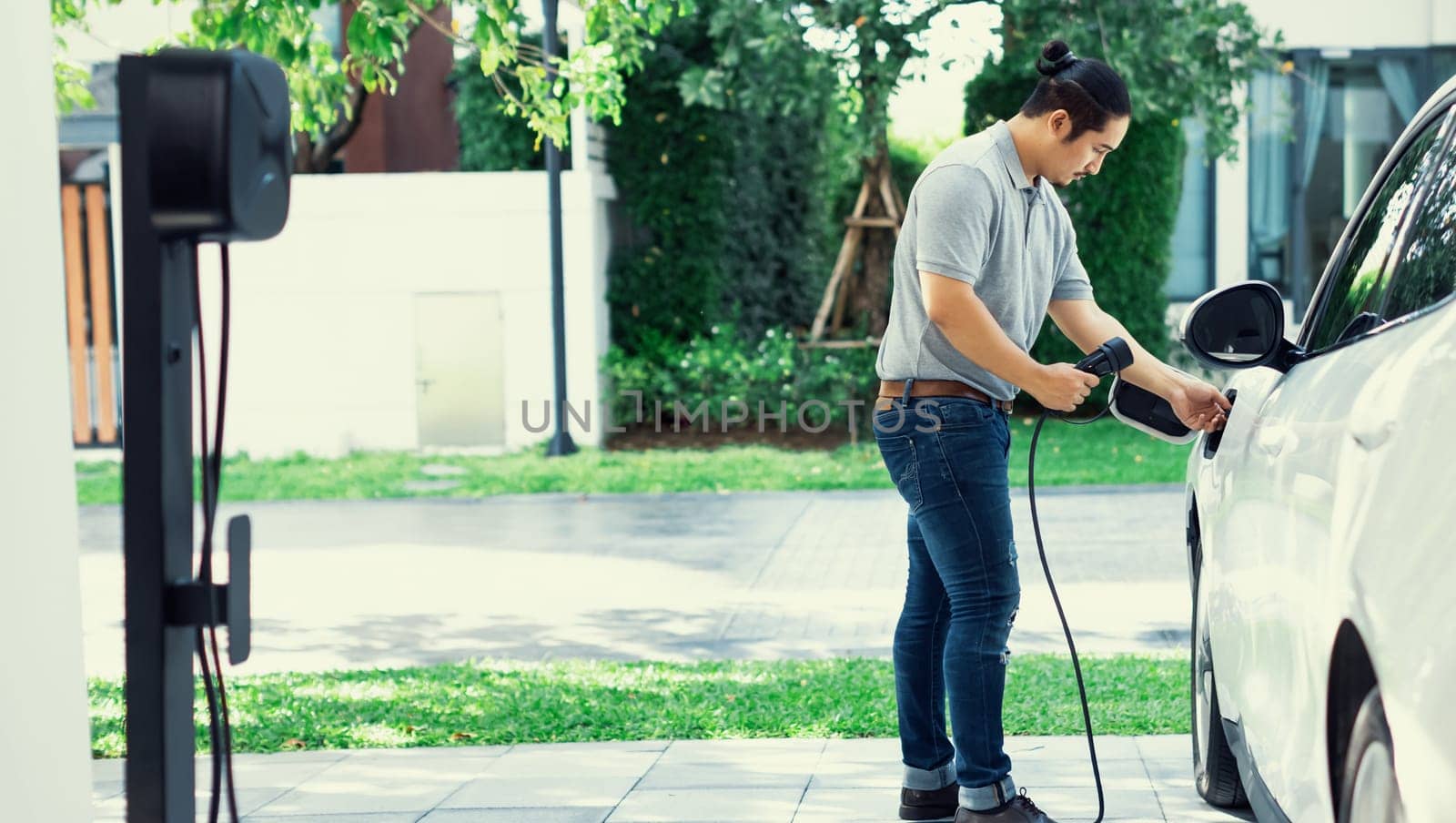 Progressive asian man recharge his EV car at home charging station. by biancoblue