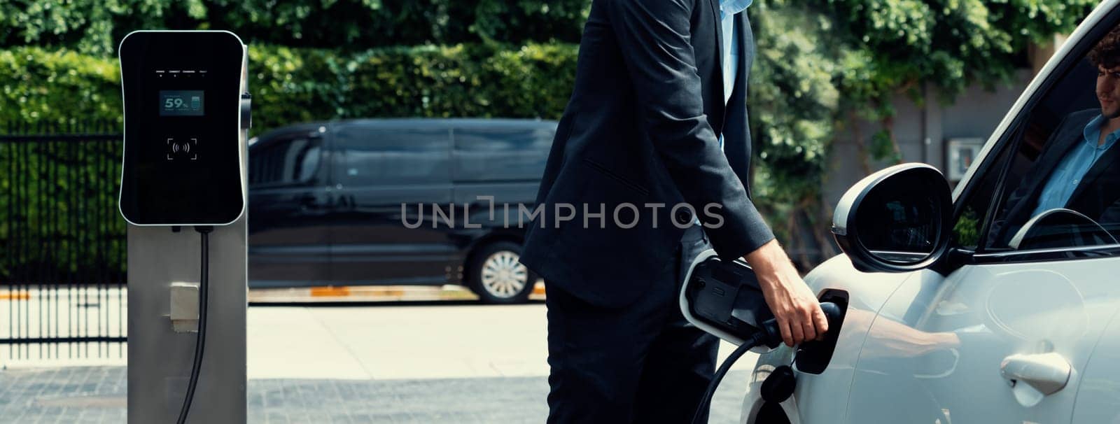 Closeup progressive black suit businessman insert EV charger plug from public charging station to his electric car. Eco friendly electric rechargeable car with zero CO2 emission.