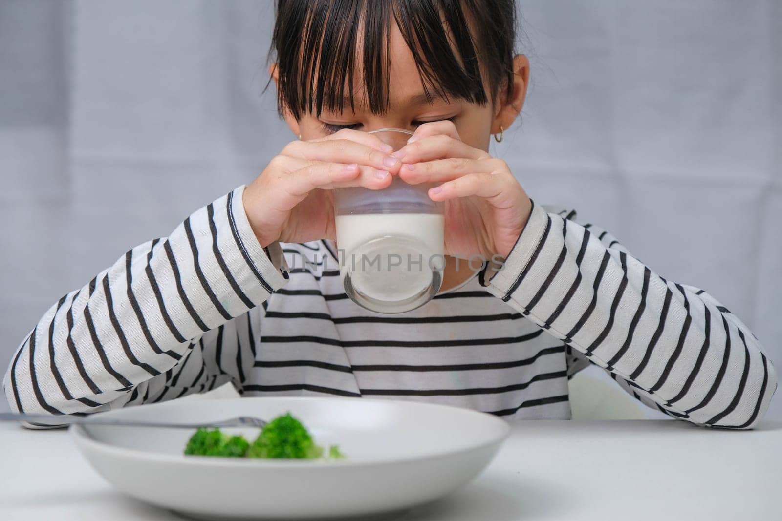 Cute Asian girl drinking a glass of milk in the morning before going to school. Little girl eats healthy vegetables and milk for her meals. Healthy food in childhood. by TEERASAK