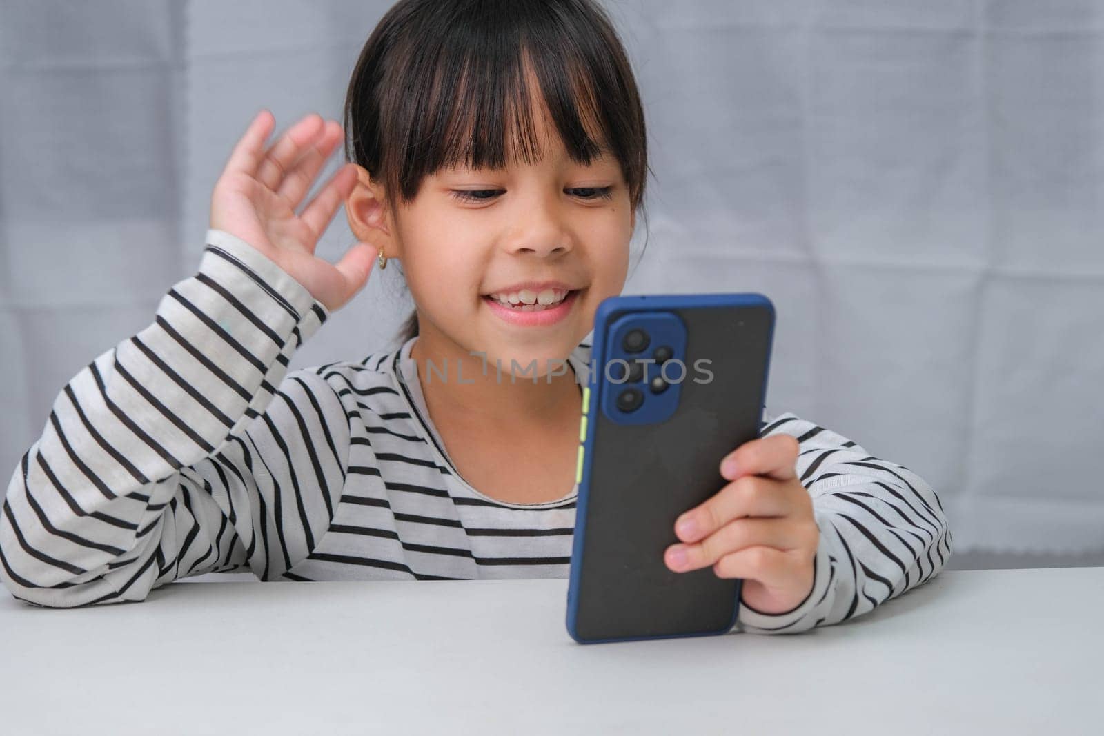 Cute little girl using smartphone and smiling while sitting at table in house. Happy Asian girl holding phone in hands, online learning or homeschooling, listening to music or playing games.