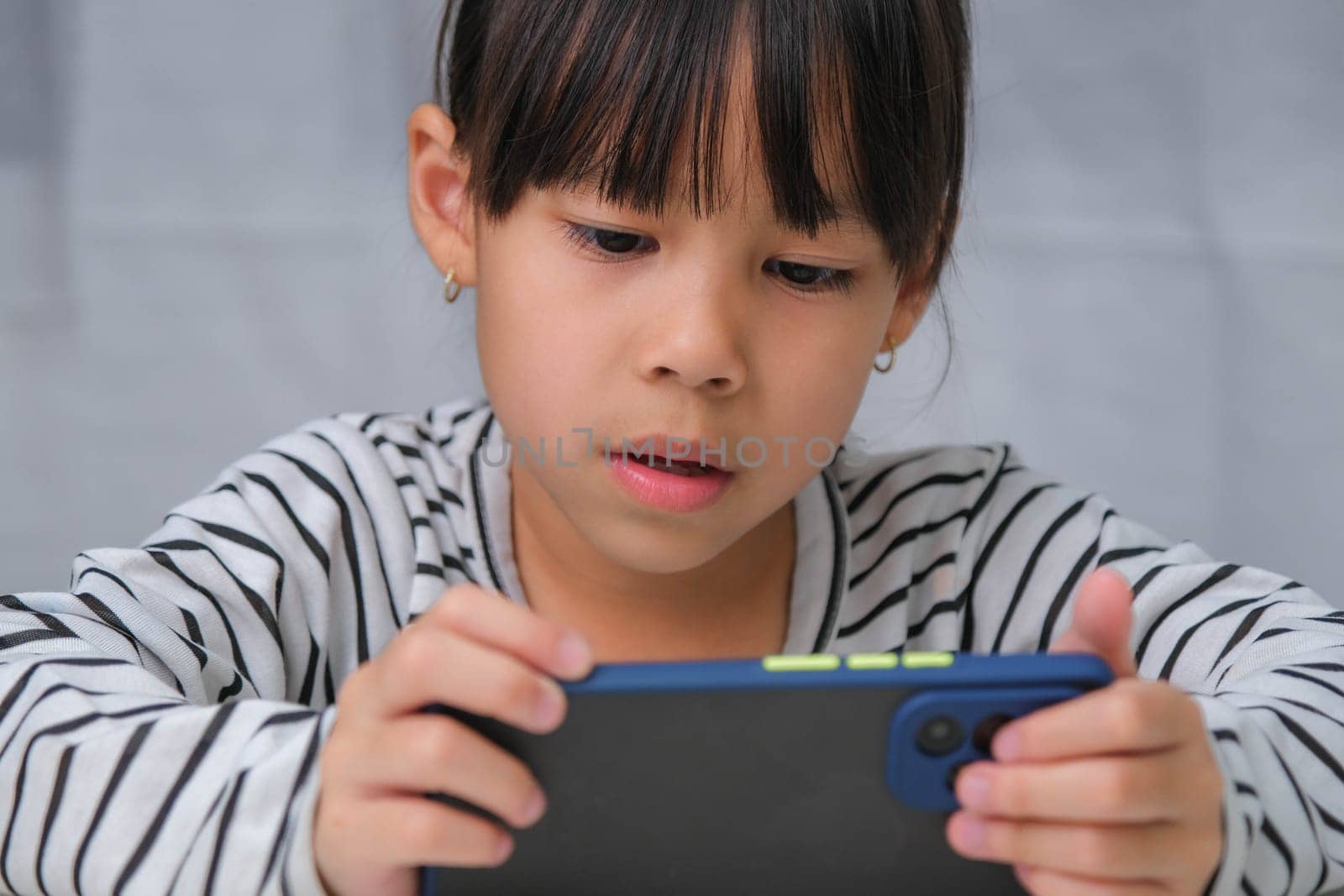 Cute little girl using smartphone and smiling while sitting at table in house. Happy Asian girl holding phone in hands, online learning or homeschooling, listening to music or playing games. by TEERASAK