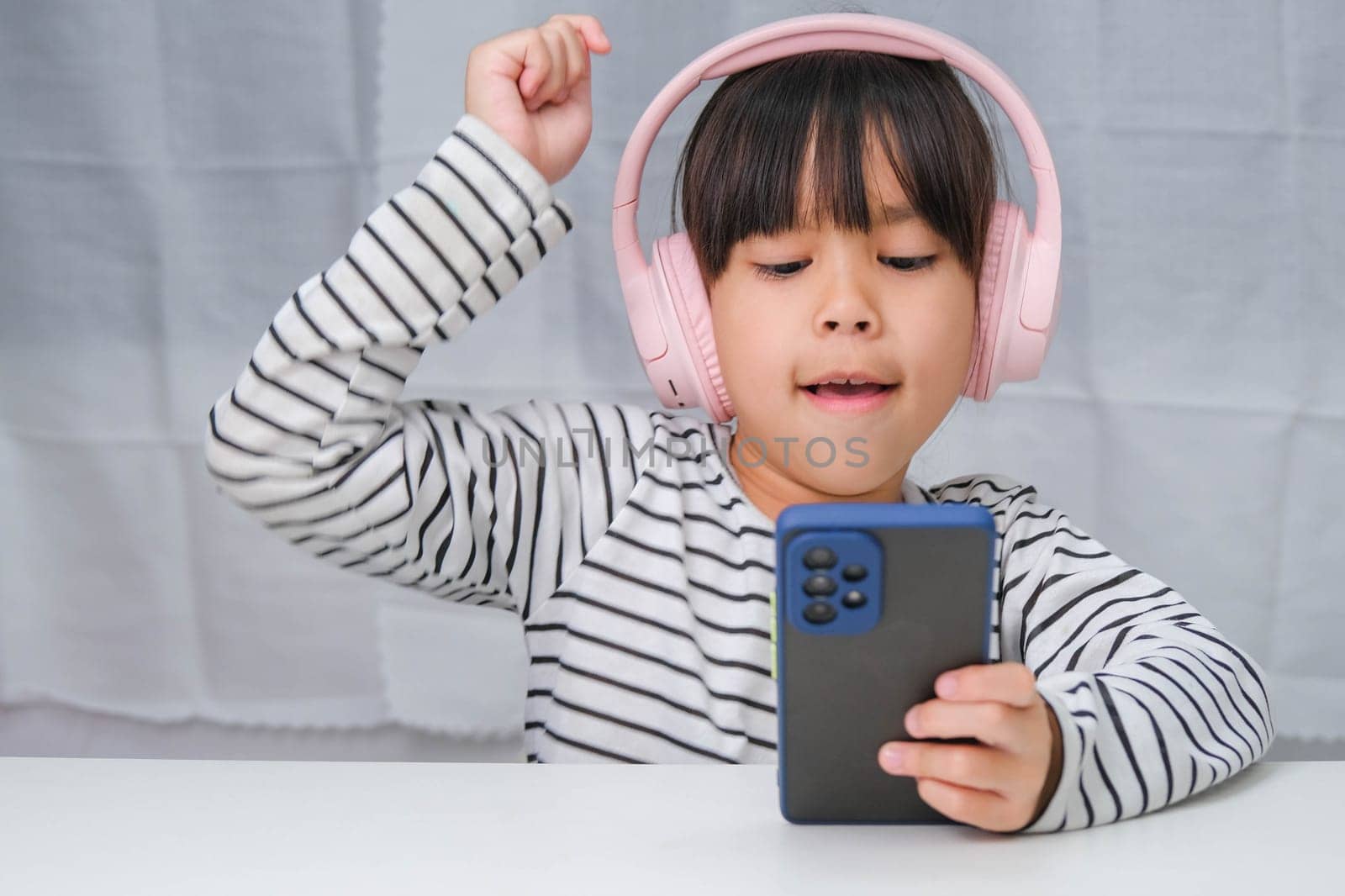 Cute elementary school girl wearing headphones holding a smartphone. Happy Asian girl studying online on smartphone or homeschooling, listening to music or playing games.