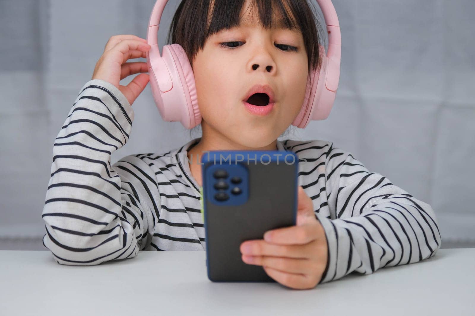 Cute elementary school girl wearing headphones holding a smartphone. Happy Asian girl studying online on smartphone or homeschooling, listening to music or playing games.